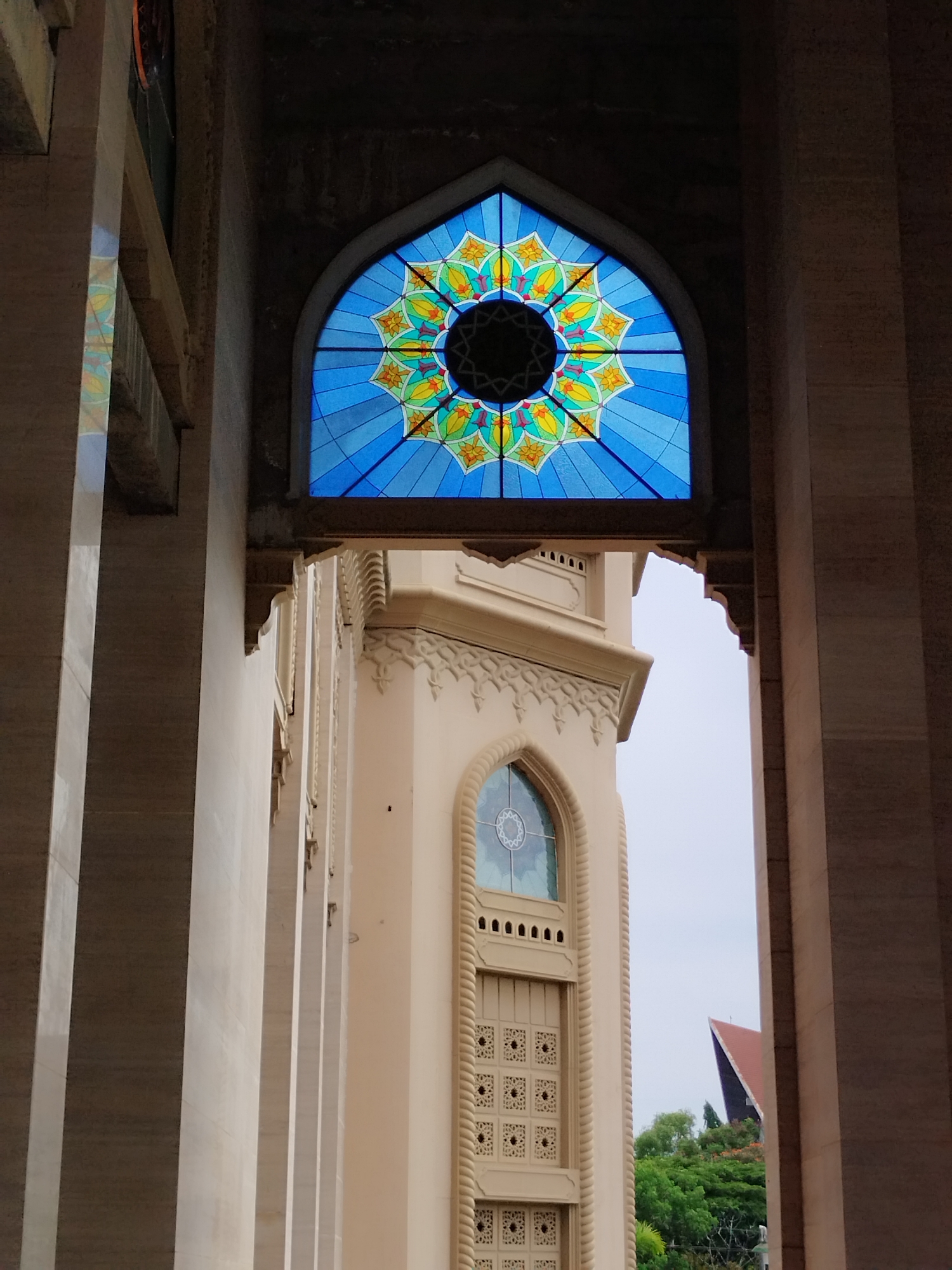 This image captures a view through the colonnade of the Islamic Center Masjid Agung Islamic Center in Lhokseumawe, Indonesia. A prominent arched stained-glass window, featuring a vibrant blue background with a geometric floral design in yellow and orange, is centered in the upper portion of the frame. Below the stained-glass window, a beige tower with decorative carvings and another smaller arched window is visible. In the distance, a red-tiled roof of a building peeks out from behind lush green trees under an overcast sky. The foreground is dominated by the large, light-colored columns of the building's architecture, casting shadows and creating a sense of depth. There are no people present in the scene.