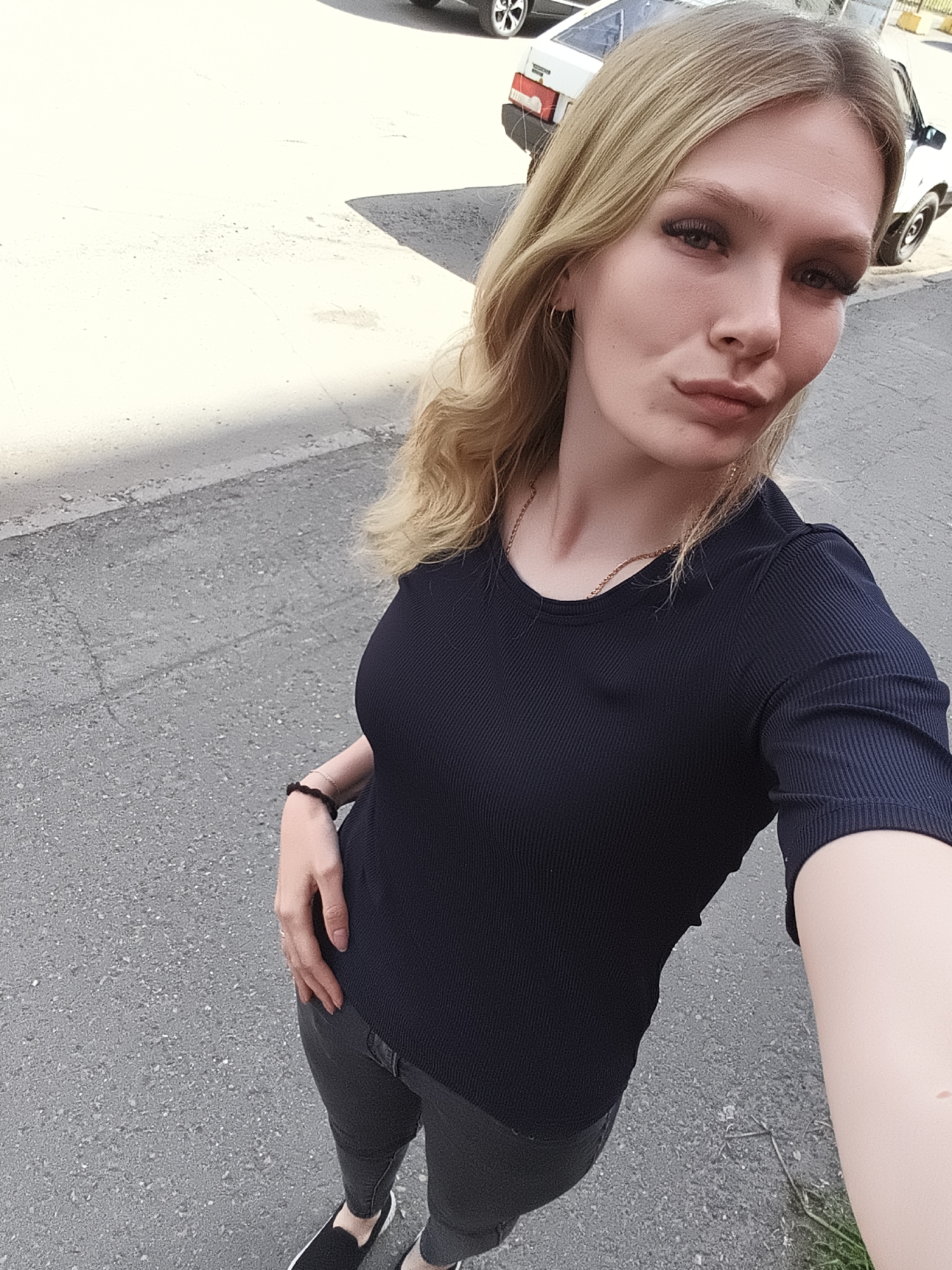 A young woman with blonde hair is taking a selfie in a city environment. She is wearing a dark blue ribbed t-shirt, dark pants, and black slip-on shoes. She has on a thin gold necklace and a black beaded bracelet on her left wrist. She is standing on a paved surface, likely a street or sidewalk. In the background, two white cars are visible. The lighting suggests it is daytime, with shadows indicating sunlight. The background details do not provide specific landmarks to pinpoint the exact location within Izhevsk, Russia.