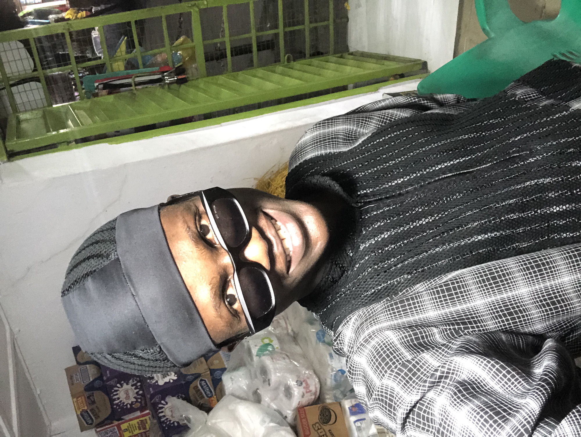 A male individual, wearing a black cap and a patterned shirt, is pictured in a room that appears to be a storage area. The individual is smiling and looking towards the camera, with dark-rimmed sunglasses obscuring their eyes. The background features stacks of various products, including boxes of what seem to be toiletries or snacks, and some rolls of toilet paper. To the right of the frame, a green metal structure, possibly shelving or a gate, is visible, with some items hanging on it. The lighting suggests an indoor setting, possibly a shop or warehouse. The presence of numerous packaged goods implies a commercial or storage environment, consistent with a location in the city of Ado, Nigeria, where small businesses and storage facilities are common.