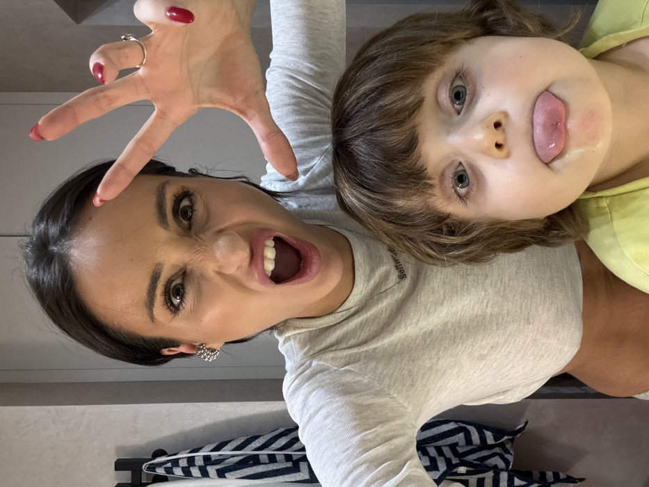 The image captures a playful moment between an adult woman and a young child, appearing as a close-up, possibly a selfie, taken from a slightly upward angle, with the subjects appearing upside down as if looking down at the camera.
The adult woman, positioned on the left side of the frame (if viewed upright), has dark hair and an expressive, exaggerated facial expression. Her eyes are wide open, looking directly at the viewer, with her eyebrows furrowed, creating a playful "monster" or "growling" look. Her mouth is open wide, revealing her teeth, in a shout or roar. She has dark mascara on her eyelashes and well-defined eyebrows. Her left hand is raised in front of her face, fingers spread wide like claws, and her fingernails are painted with dark red polish. She wears a silver-toned ring on her left ring finger and a decorative, possibly floral or cluster-style, silver earring in her left ear. She is wearing a light grey, long-sleeved top. On the left side of her chest, faint text is visible, which appears to read "Selfmade."
Next to her, on the right side of the frame, is a young child. The child has light brown, shaggy hair with bangs and bright blue eyes that also look directly at the viewer. The child's facial expression is equally playful, with their tongue sticking out of their mouth. The child has fair skin and somewhat rosy cheeks, suggesting youth. They are wearing a light yellow or greenish top.
The two subjects are positioned very close together, creating a sense of intimacy and shared fun. Their playful expressions suggest they are engaged in a game or creating a silly memory.
The background is simple and out of focus. Behind the woman, a light-colored wall is visible. Below the subjects, a dark, possibly metal, bar or rack is partially visible, with a striped towel featuring a navy blue and white chevron or zig-zag pattern hanging on it. This detail suggests an indoor setting, possibly a bathroom or a casual living space within a home. The lighting is bright and even, indicating an indoor environment, likely during the daytime or in a well-lit room. The overall mood of the image is joyful, spontaneous, and affectionate.
