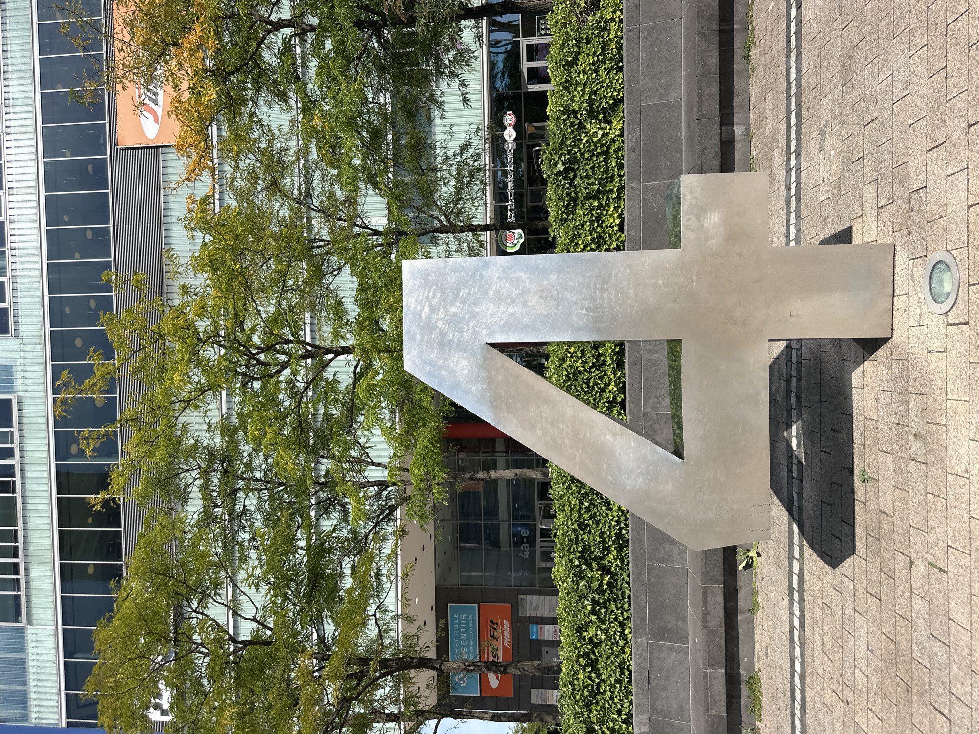 The image depicts a large, freestanding, metallic number "4" positioned on a paved outdoor area. Behind the number, a modern building with a glass facade is visible, featuring horizontal window bands and what appears to be a green, textured siding. Trees with green foliage are situated between the number and the building. A dense hedge runs horizontally across the mid-ground, separating the paved area from the building's lower level, which shows storefronts with signage.
Based on the visible signage, specifically "HOCHSCHULE GENIUS" (University of Applied Sciences) and "S' Fit Premium", and the architectural style, this location is identifiable as the **Cologne University of Applied Sciences (TH Köln)** campus at **Claudiusstraße 1, 50674 Köln, Germany**. The "4" sculpture is a prominent feature on this campus, often seen as an artistic element or address marker. No people are present in the immediate foreground or mid-ground. The lighting suggests a daytime scene, with shadows cast by the sculpture. The pavement is made of interlocking rectangular stones.