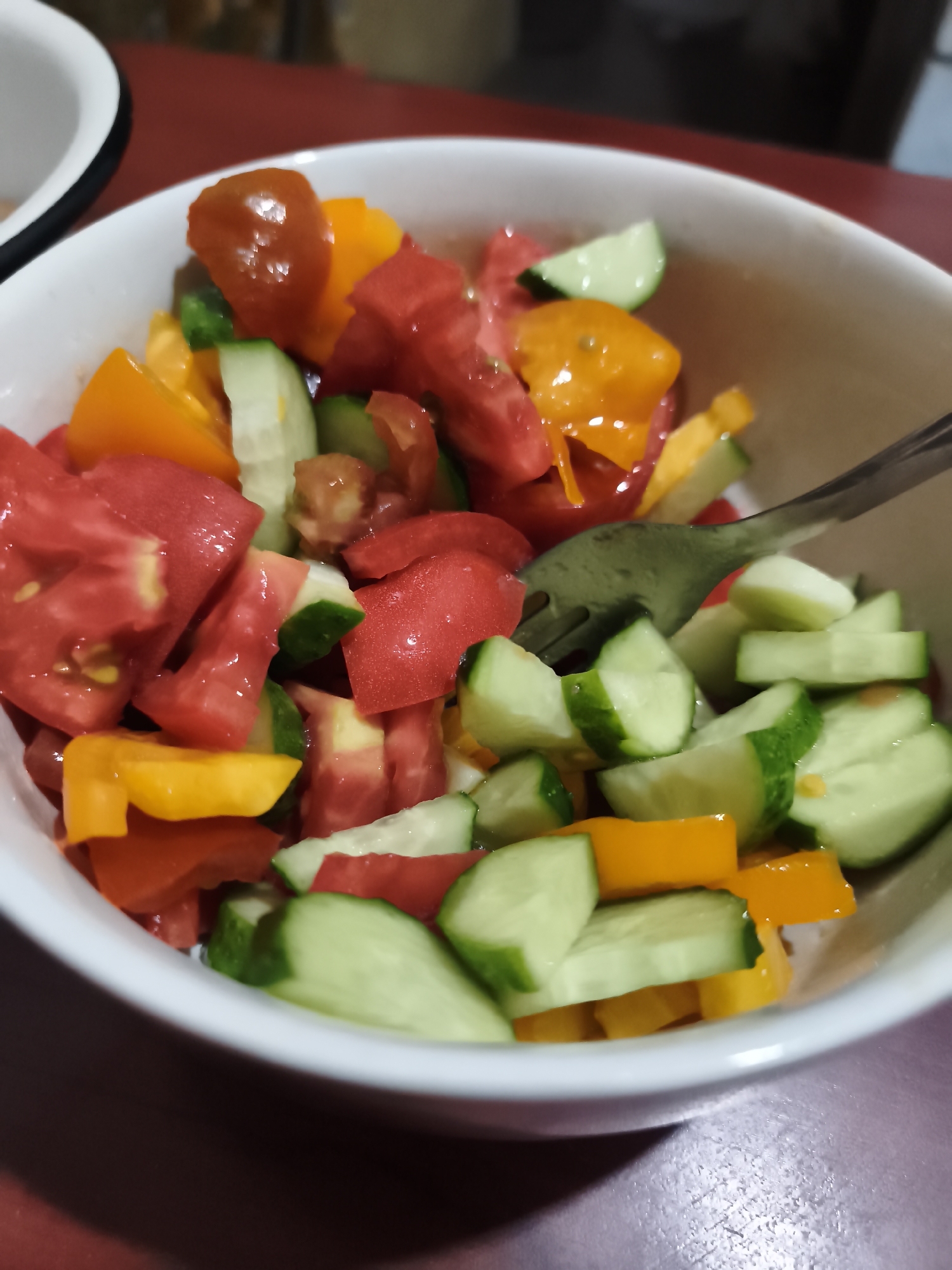 A close-up, eye-level shot shows a white bowl filled with a chopped tomato and cucumber salad. The salad contains red tomatoes, orange bell peppers, and green cucumbers, all cut into bite-sized pieces. A fork is partially visible in the right side of the bowl, with some salad ingredients on its tines. The bowl is set on a dark red surface, possibly a tablecloth or countertop. The background is blurred, but suggests an indoor setting. The scene is devoid of any recognizable landmarks or specific details that would indicate its location in Sokolivske, Ukraine.