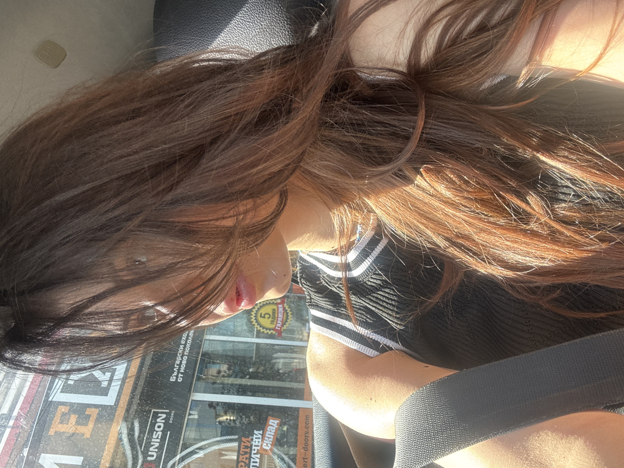A medium shot captures a woman in a car, with her dark brown hair partially obscuring her face. She is wearing a black sleeveless top with white stripes on the straps. A grey seatbelt is visible across her chest. Through the car window, a storefront with signs is visible, including text that appears to be in Cyrillic script and the word "UNISON". The sunlight is strong, casting shadows and highlighting the texture of her hair and skin. The background suggests an urban environment, likely a street in Sofia, Bulgaria, given the Cyrillic text.