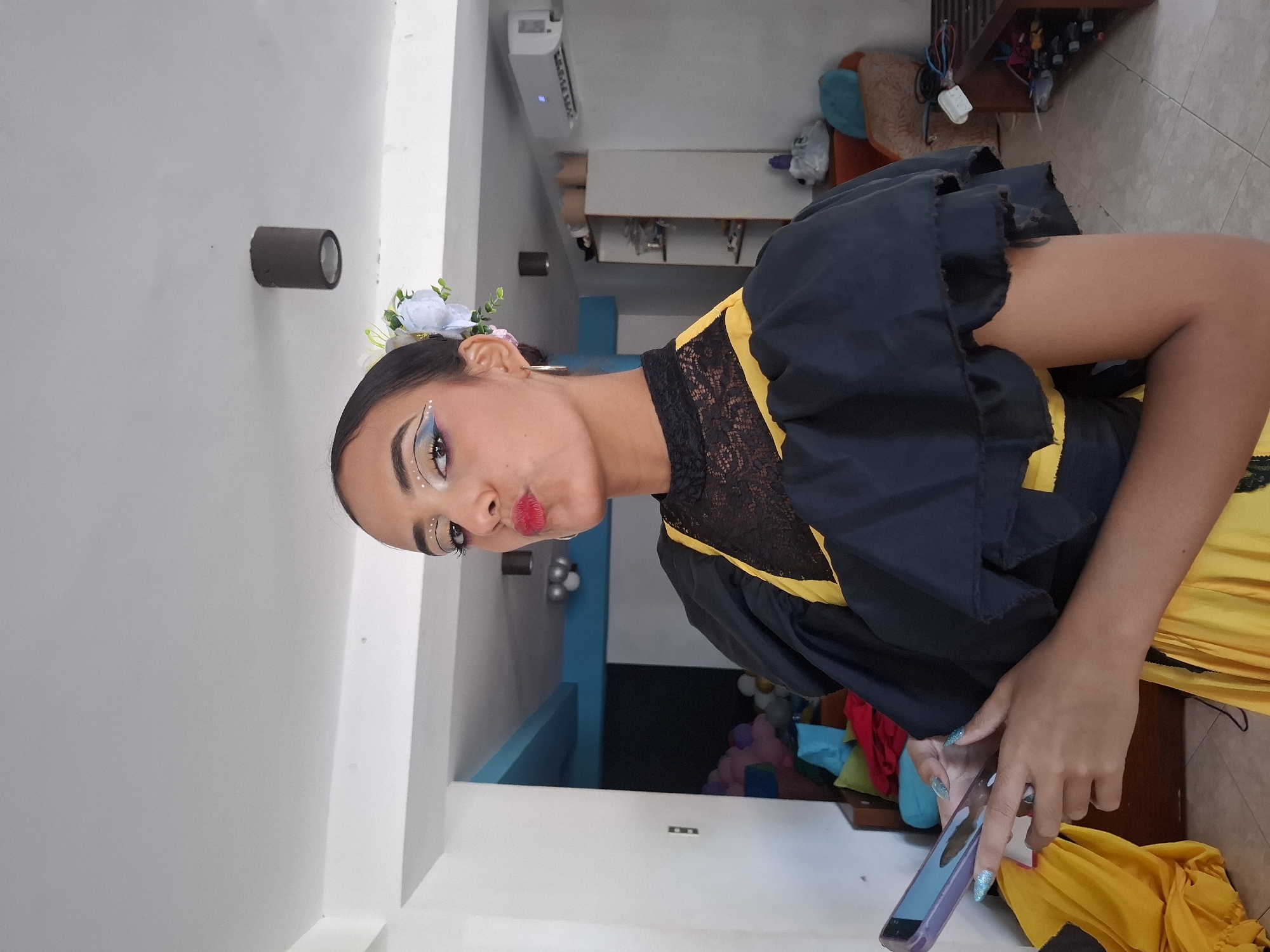 A woman is shown indoors, presumably in a home or apartment, in Playa El Agua, Venezuela. She is wearing a traditional black and yellow dress with a black lace collar and has flowers in her hair. She is holding a smartphone in her hands, seemingly interacting with it. The background includes white walls, a light fixture, and glimpses of other furniture and decor, including balloons and what appear to be clothing items.