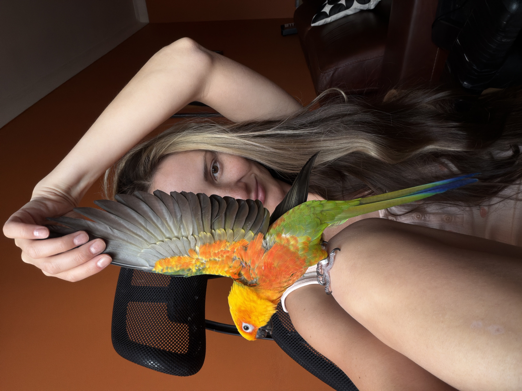 A young woman with long, wavy blonde and brown hair is lying down on a black office chair, her arm bent and resting above her head. She is looking towards the camera with a slight smile, her eyes brown and her face lit by natural light. A colorful sun conure parrot, predominantly orange and yellow with green and blue wing feathers, is perched on her arm. The parrot's wings are spread, displaying its vibrant plumage. Its tiny feet are visible gripping the woman's skin, and it wears a silver band on one leg. The background is a warm, orange-toned wall, suggesting an indoor setting. To the upper right, a glimpse of a brown leather couch and a black object, possibly part of a chair or furniture, can be seen. The overall atmosphere is intimate and relaxed, hinting at a quiet moment shared between the woman and her pet. There is no visible text in the image. The lighting suggests it might be daytime, likely indoors.