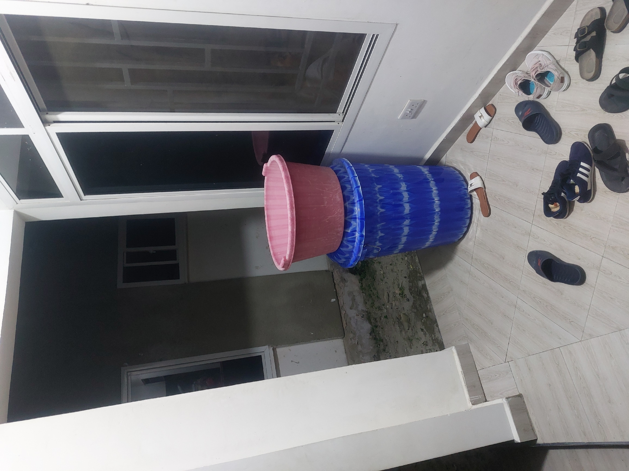 The image shows an interior space with several objects. A stack of plastic tubs, one pink and the others blue, sits near a window. Adjacent to the tubs are various pairs of shoes and sandals scattered on the tiled floor. The room appears to be a veranda or porch, with the windows looking out onto an exterior space. Based on the environment and the typical style of architecture and domestic scenes, this photo could be in Dar es-Salaam, Tanzania.