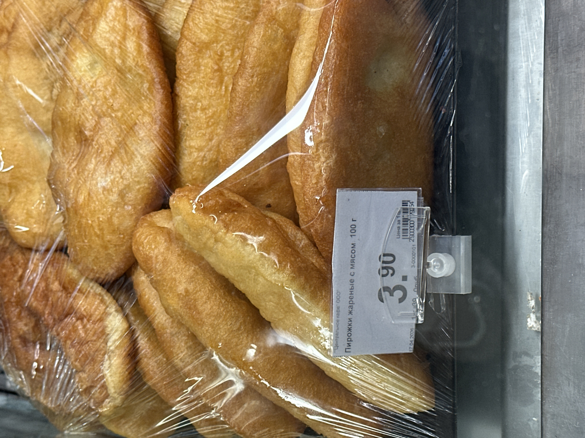 A close-up, eye-level shot shows a display of fried pastries, likely in a bakery or grocery store in Minsk, Belarus. The pastries, golden brown and wrapped in clear plastic, are stacked in multiple rows. A price tag is attached to the front of the display, showing a price of 3.90 and the text "Пирожки жареные с мясом, 100 г" (fried pies with meat, 100g). The background is out of focus, but appears to be a store shelf or refrigerated display case. The lighting is bright, highlighting the texture of the pastries.