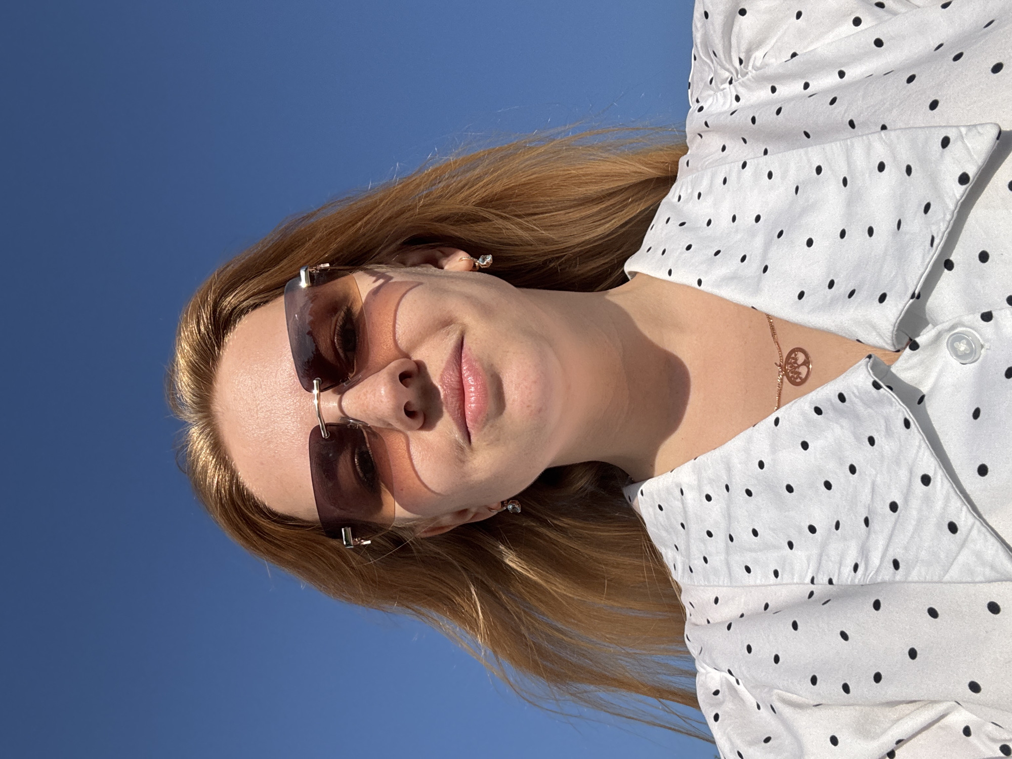 A woman with blond hair and sunglasses stands against a clear blue sky. She wears a white polka-dot shirt and a gold necklace. The location is Rusanovo, Russia.