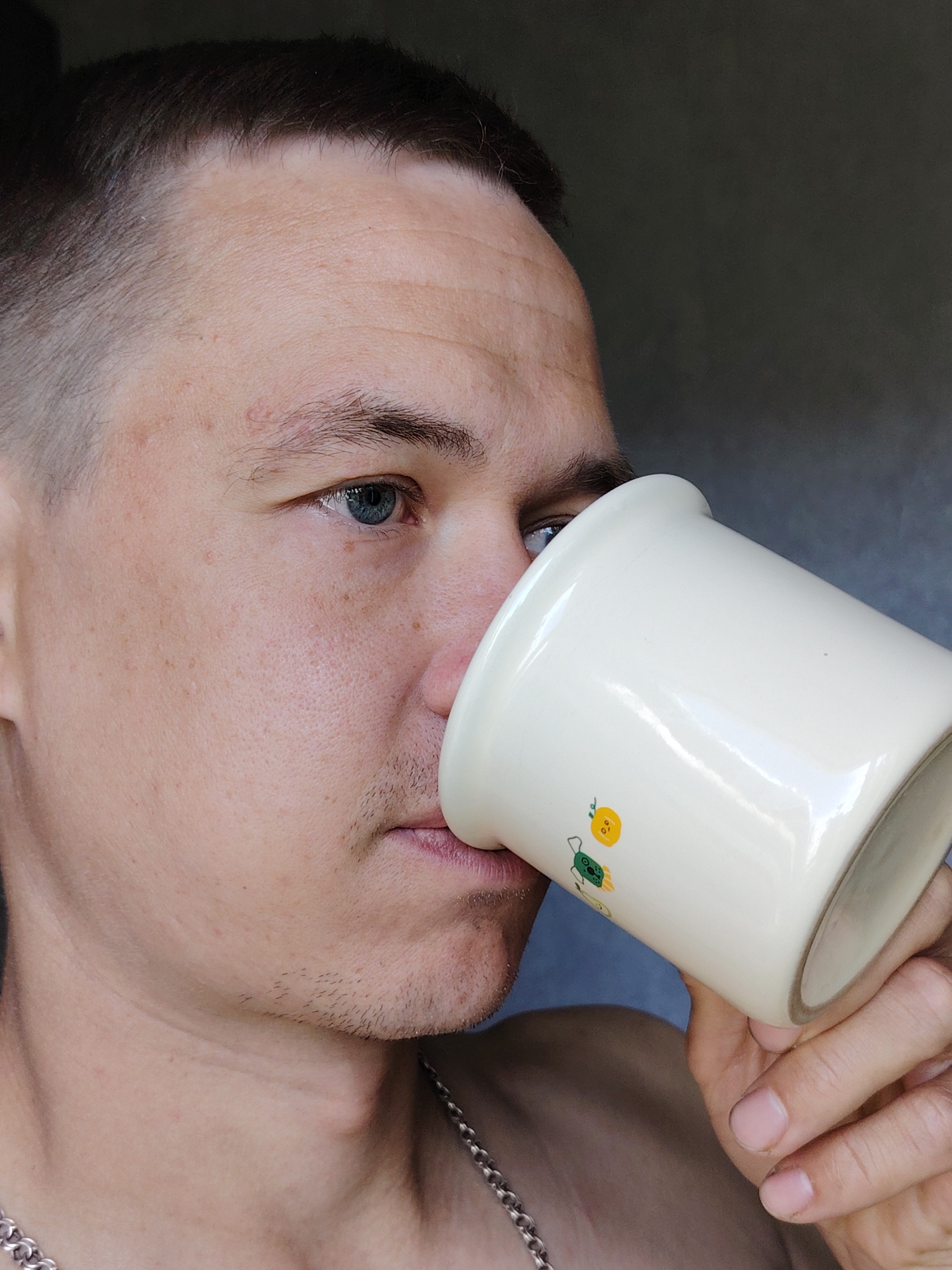 A close-up, medium shot depicts a fair-skinned man drinking from a white mug. He has short, dark brown hair and blue eyes. Some facial hair is visible on his chin. He is wearing a silver chain necklace. The mug is decorated with small cartoon-like drawings, including what appears to be an orange and a turtle. The background is out of focus, but appears to be a neutral gray color. There is no visible indication that the image was taken in Chaikovsky, Russia.