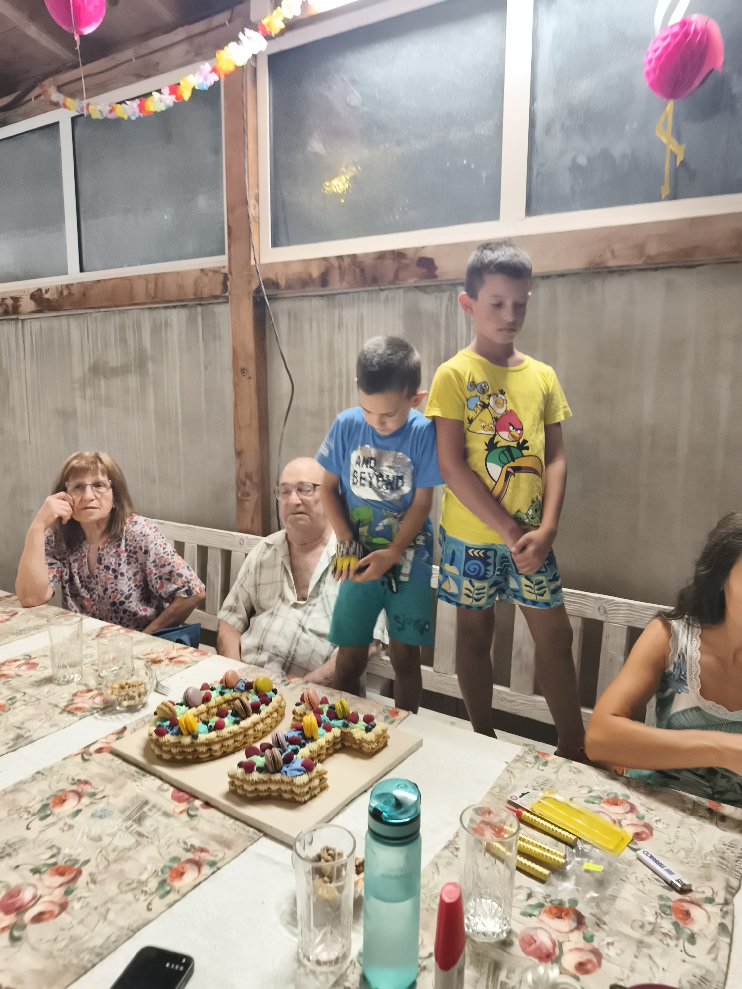A group of people, likely a family, are gathered around a table set up outdoors, possibly in a backyard or patio area. The scene appears to be a celebration, indicated by the presence of a decorative cake shaped like a number, possibly a birthday cake.
The people present include an older couple sitting at the table. The woman has brown hair and is wearing glasses and a patterned blouse. The man, also wearing glasses, is in a plaid shirt. Two young boys are standing near the table; one is wearing a blue t-shirt and shorts, and the other is in a yellow t-shirt and patterned shorts, with a design of birds on his shirt. Another individual, a woman, is partially visible on the right side of the image, wearing a white and blue top.
The table is covered with a floral-patterned tablecloth. On the table are several glasses, a water bottle, and the aforementioned cake, which is decorated with macarons, berries, and what appear to be edible flowers or frosting details. There are also some small nuts in a glass bowl and what looks like a mobile phone. Towards the right side of the table, there are some yellow objects that resemble sparklers or party poppers, along with a red item that might be lipstick or a marker.
The background shows a concrete wall and wooden beams, suggesting an outdoor structure like a pergola or a covered patio. Decorations are visible in the upper part of the image, including colorful paper garlands and a pink balloon. The overall lighting suggests it is either dusk or evening, or the area is dimly lit.
The exact location in Stamboliyski, Bulgaria, is not specified within the image itself, but the context suggests a residential outdoor setting for a family gathering.
