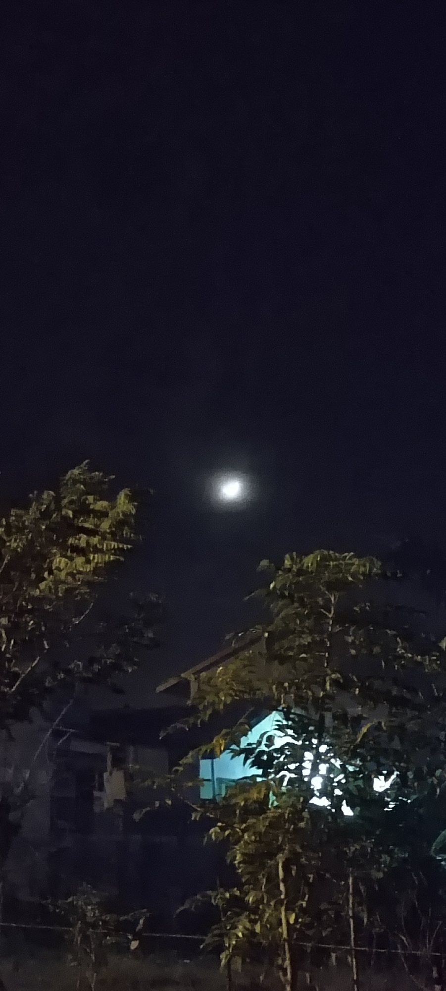 The image depicts a nighttime scene in Lhokseumawe, Indonesia. A bright full moon is visible in the dark sky. In the foreground, lush green trees with dense foliage partially obscure a building with a light blue wall and a steeply pitched roof. The building appears to be illuminated from within, casting a bright glow through the leaves. In the background, a darker structure, possibly a house, is faintly visible behind the trees. The overall impression is a quiet, residential area under the glow of the moon.