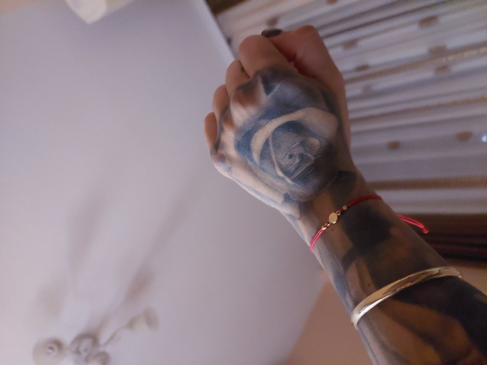 The image shows a person's hand and forearm, heavily tattooed with dark ink. A blue rose tattoo is prominent on the back of the hand. The person is wearing two bracelets on the wrist: a thin red string bracelet with gold beads and a simple gold cuff bracelet. The background is blurry, suggesting an indoor setting with a white ceiling and what appears to be a wall or surface with horizontal lines and decorative elements. There is no specific evidence within the image to identify the location as Plovdiv, Bulgaria.