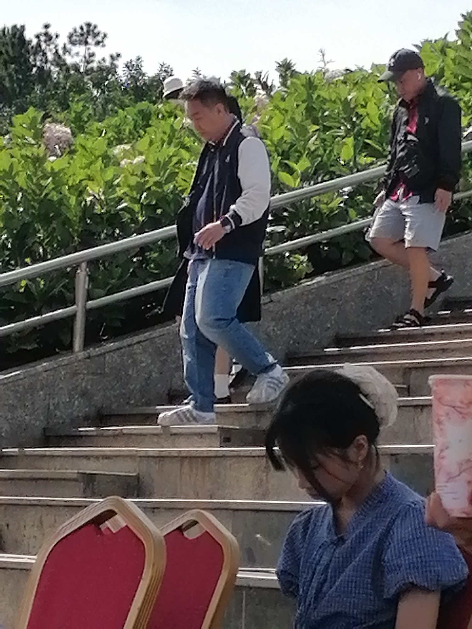 In Da Lat, Vietnam, on a bright day, three individuals are captured on an outdoor staircase surrounded by lush green foliage. In the foreground, a young woman with dark hair, wearing a blue patterned shirt, is seated with her head bowed. She is partially obscured by the edge of a red chair with a wooden frame. Two men are ascending the stairs. The man closer to the viewer, wearing a dark jacket with white sleeves, blue jeans, and white athletic shoes, is walking down the stairs. To his right, another man in a black jacket, plaid shirt, light-colored shorts, and a baseball cap is walking up the stairs. A silver railing runs along the side of the stairs. The environment suggests a park or a scenic viewpoint, common in Da Lat's hilly terrain.