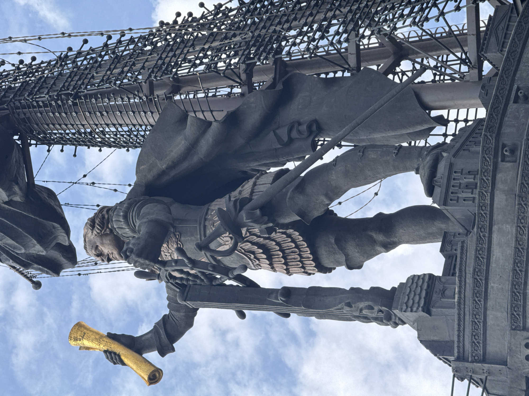 An outdoor, low-angle, medium shot shows a monumental statue of a man dressed in a cape and what appears to be armor with a scalloped skirt. The statue is made of a dark metal, possibly bronze, and stands against a cloudy blue sky. The man holds a golden, rolled-up scroll in his raised left hand and grips a ship's wheel with both hands. His right hand is positioned on the ship's wheel, while his left hand is slightly above it. His head is turned to his left, and he appears to be looking forward.
Behind the statue, a tall, dark metal structure that resembles a ship's mast with rigging and netting rises into the sky. The base of the statue is elaborate, with architectural details that suggest a ship's deck or platform, including tiered levels and decorative carvings. The overall scene conveys a sense of maritime exploration or leadership. The location is identified as Unknown, Unknown.