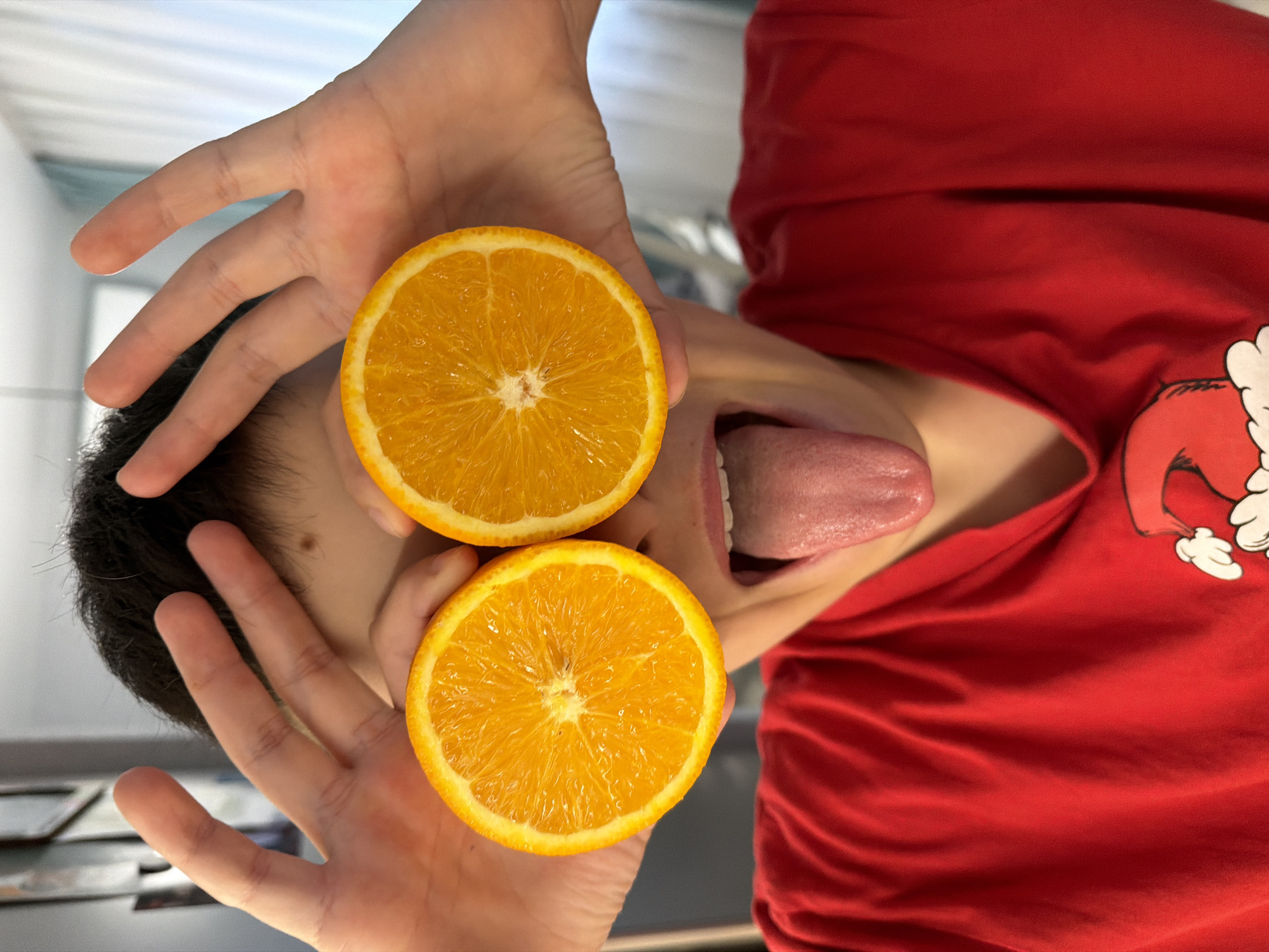 A person wearing a red shirt with a white graphic is holding two orange halves in front of their face, with their tongue sticking out between the orange slices. The person's hands are visible, framing the orange halves. The background is blurred, showing a glimpse of an interior space. There is no indication of this being in Votkinsk, Russia.
