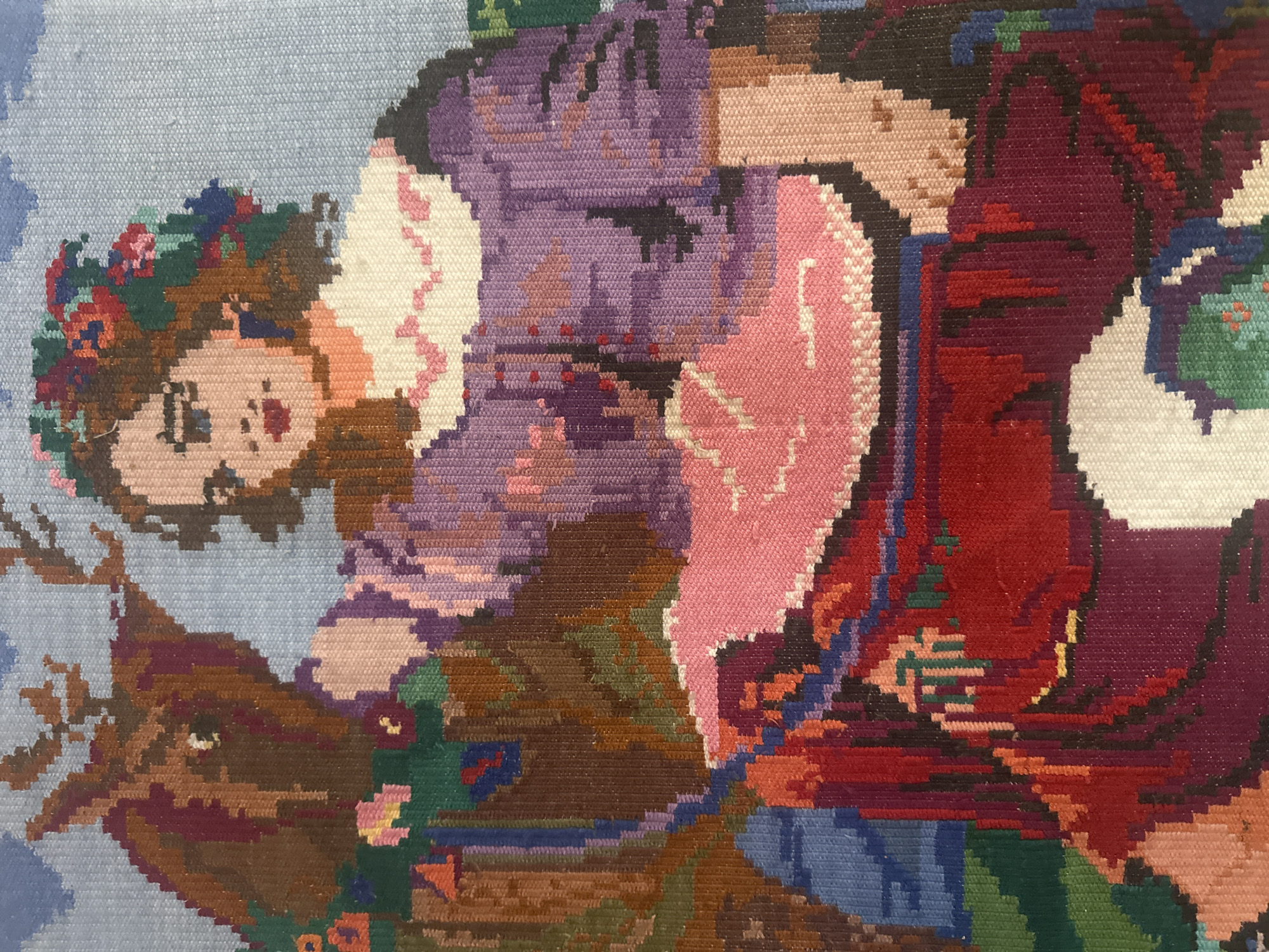 This is a close-up, horizontally oriented image of a woven tapestry depicting a person. The style is pixelated, characteristic of needlepoint or cross-stitch embroidery. The central subject appears to be a young girl with a light complexion, large dark eyes, and rosy cheeks. She wears a crown or headdress adorned with colorful flowers and greenery in shades of red, pink, green, and blue. Her hair is brown. She is positioned in the foreground, facing slightly to the right.
Behind and around her, the tapestry is filled with a riot of colors. To her right, a figure in a large, voluminous garment dominates the middle ground. This figure is dressed in shades of purple, burgundy, and red, with accents of pink and white on what appears to be a collar or headdress. The garment has intricate patterns and folds, rendered in the characteristic stitch-by-stitch style of tapestry.
To the left of the girl, partially visible, is what seems to be the head and neck of a brown animal, possibly a horse or a deer, with dark eyes and some antler-like protrusions. The background is a soft, light blue with some cloud-like formations. Scattered throughout the tapestry are floral motifs and abstract shapes in various colors, including green, brown, and blue.
The overall impression is that of a folk art depiction, possibly a scene from a story or a festive celebration. The emotions conveyed are somewhat ambiguous due to the stylized nature of the artwork, but the girl's expression could be interpreted as calm or even a little sad, while the vibrant colors suggest a lively setting. There is no visible text in the image. The location context of Пярэжырскі сельскі Савет, Belarus, suggests a possible origin and cultural context for this tapestry, hinting at traditional Belarusian artistic motifs.