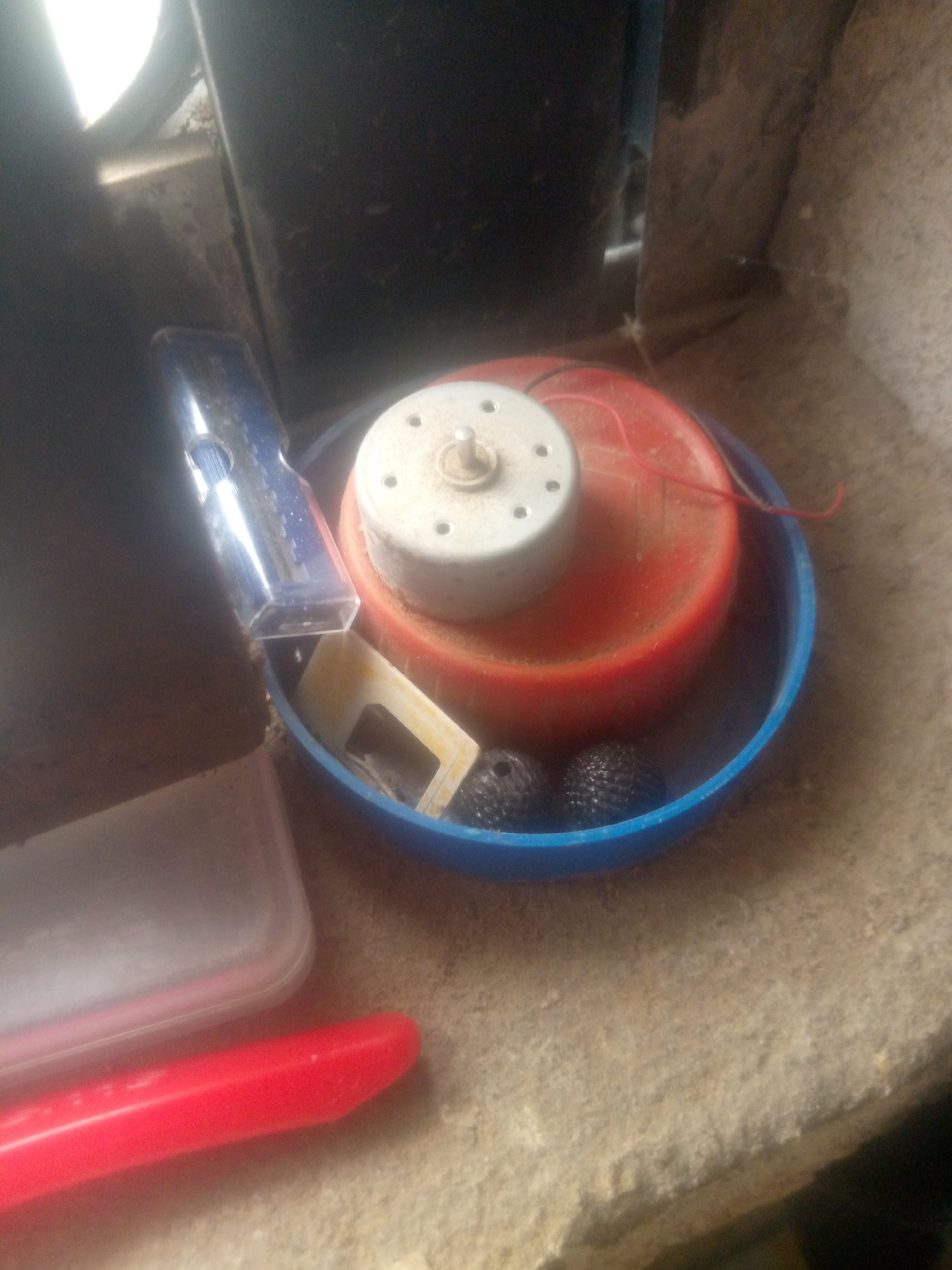 A blue plastic basin holds a disassembled, possibly homemade, motorized device. In the center of the basin is a red cylindrical object with a white motor assembly on top, featuring a central shaft and several screw holes. A thin red wire extends from the motor. To the left of the motor assembly, a blue transparent plastic tool with a silver metallic tip is partially submerged in the basin. Below the motor assembly and to the right are two black textured spheres. To the left of the basin, a clear plastic container with a pinkish-red lid sits on a textured, beige surface. In the foreground, a bright red object, possibly a tool handle, lies horizontally. The background is dark and indistinct, suggesting an indoor or shadowed outdoor setting. The overall scene appears to be an assortment of discarded or unassembled electronic components and tools.