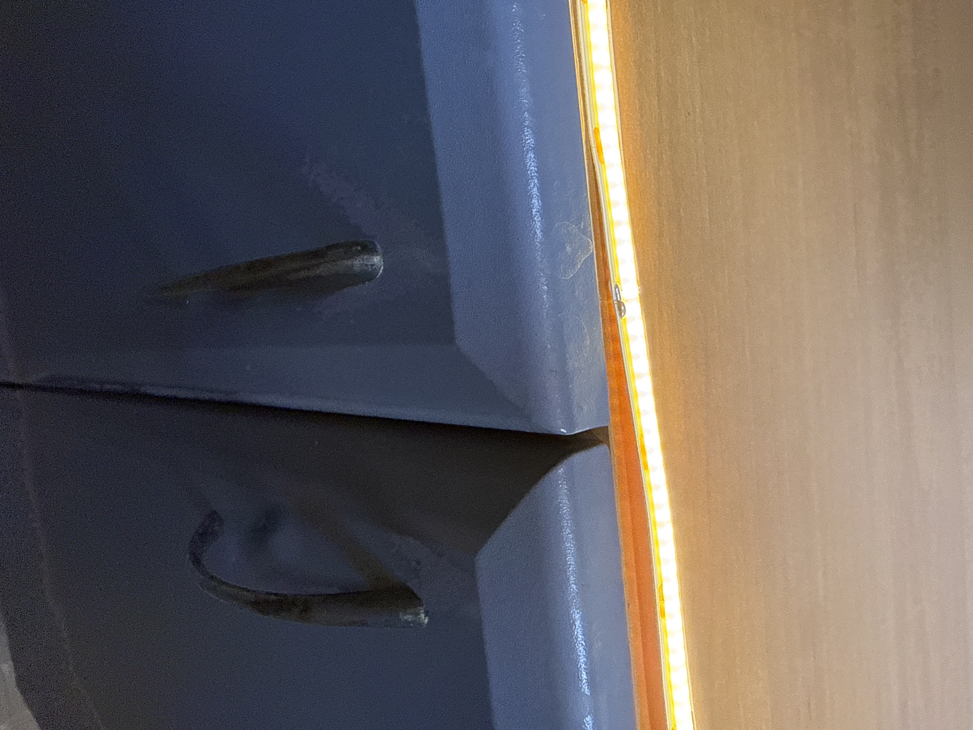 The image shows a close-up, eye-level view of two dark gray, curved indentations against a darker background. Both indentations are roughly the same size and shape, resembling handles or grips. The left indentation has a pronounced curve, while the right one is straighter. The surface surrounding these indentations appears to be a flat, textured material. Below these indentations, a bright, linear light source with many small, individual bulbs illuminates a wooden surface. The lighting casts shadows and highlights on the indentations, suggesting they are part of a larger structure or object. The scene is devoid of people or any overt activity. The overall impression is one of detail and texture rather than a broad environmental context.