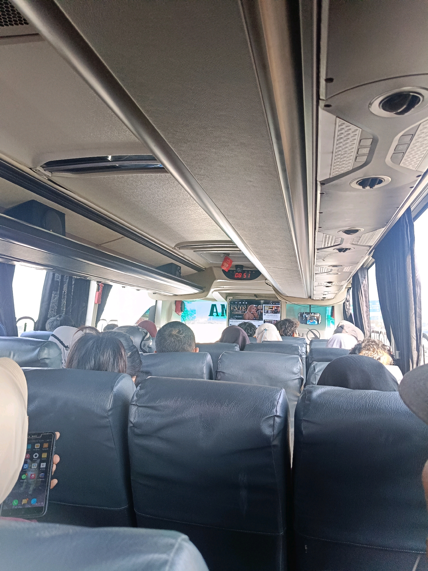 The image shows the interior of a bus, likely in Bungurasih, Indonesia. The perspective is from the back of a seat looking forward. Passengers are seated in rows of dark-colored bus seats. The ceiling of the bus features overhead compartments and air vents. A digital clock displaying "08 51" is visible above a screen showing what appears to be entertainment or a travel announcement. One passenger in the foreground is holding up a smartphone. The overall scene depicts a typical moment of travel inside a bus.