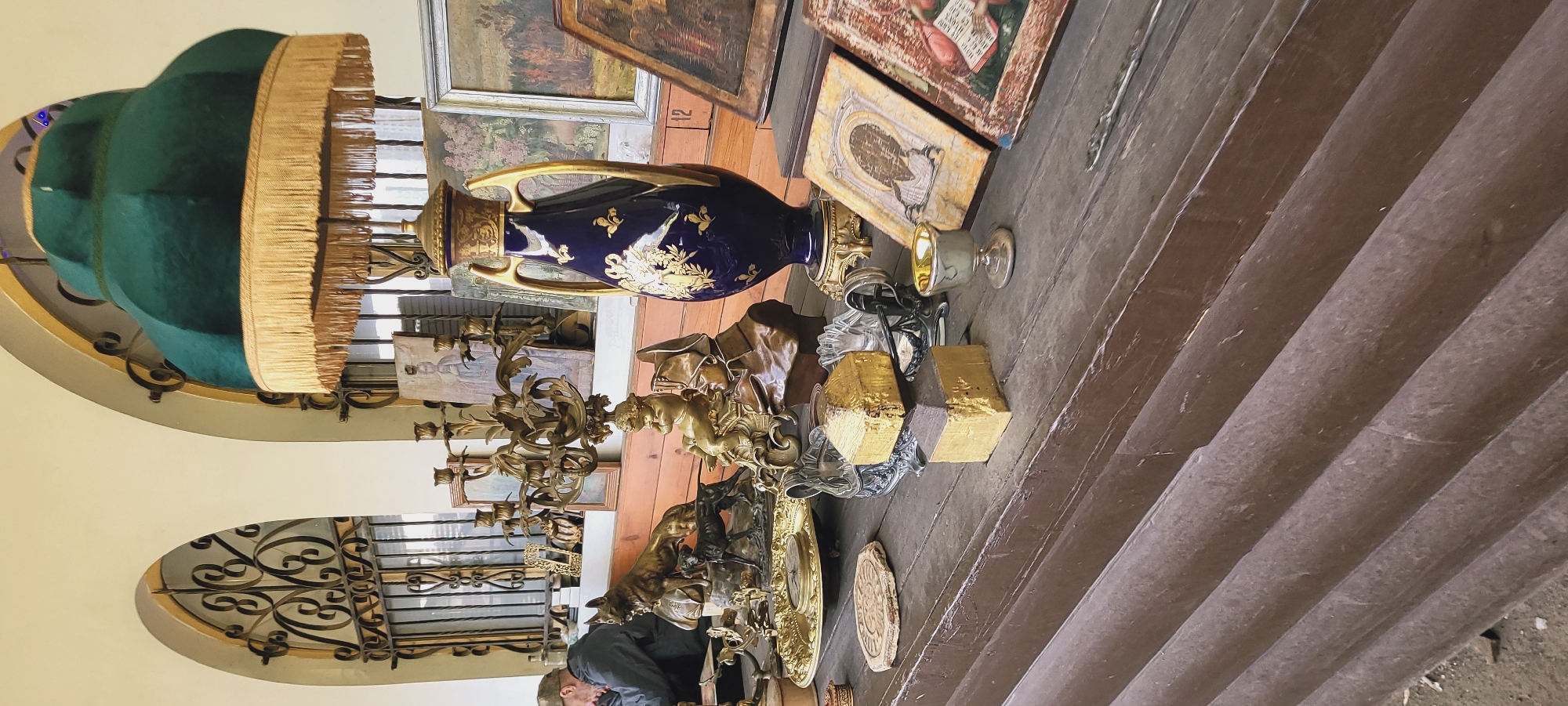 Here's a description of the image:
**Scene:** An indoor setting cluttered with antiques and artifacts, likely in a shop or storage area.
**Objects:** There's a large green-shaded lamp with a fringe, a dark blue vase with gold accents, several paintings, candelabras, figurines of people and animals (possibly bronze), decorative objects, a small goblet. Some objects appear to be gold-plated. There's also a wooden surface that looks like stairs, where items are arranged.
**People:** There appears to be a person at the bottom of the image, facing toward the left of the image.
**Environment:** The background includes a wall with archways, windows with decorative metal grilles, and other items in the background. The lighting is mixed, with some objects highlighted and others in shadow.
**Location:** Unknown city, unknown.