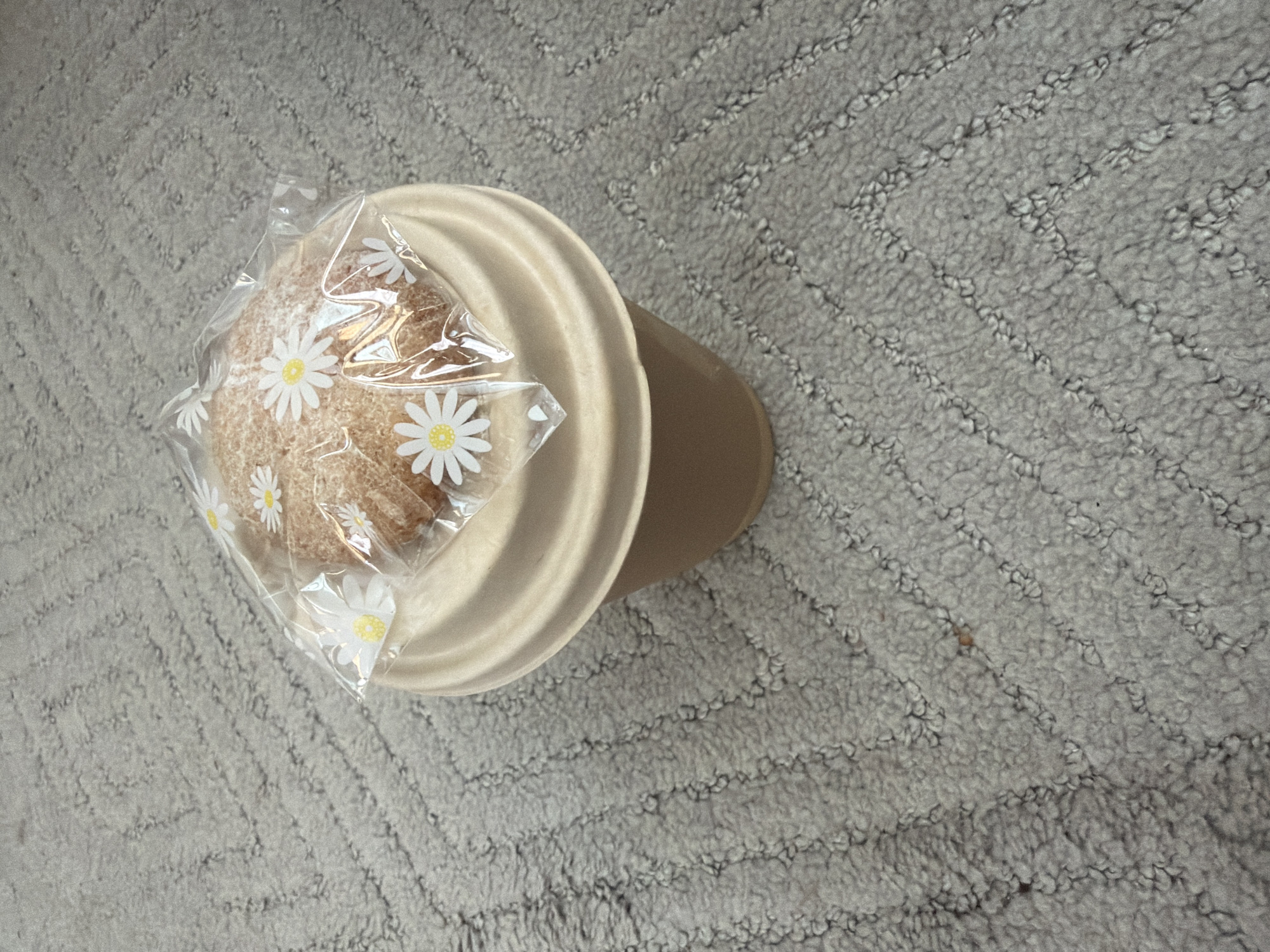 A top-down, eye-level photograph depicts a muffin in a clear plastic wrapper adorned with white daisy flowers, sitting atop a pale tan paper cup and lid. The objects rest on a gray carpet with a repeating geometric pattern. The location is undetermined as there are no specific indicators to pinpoint Ungasan, Indonesia.