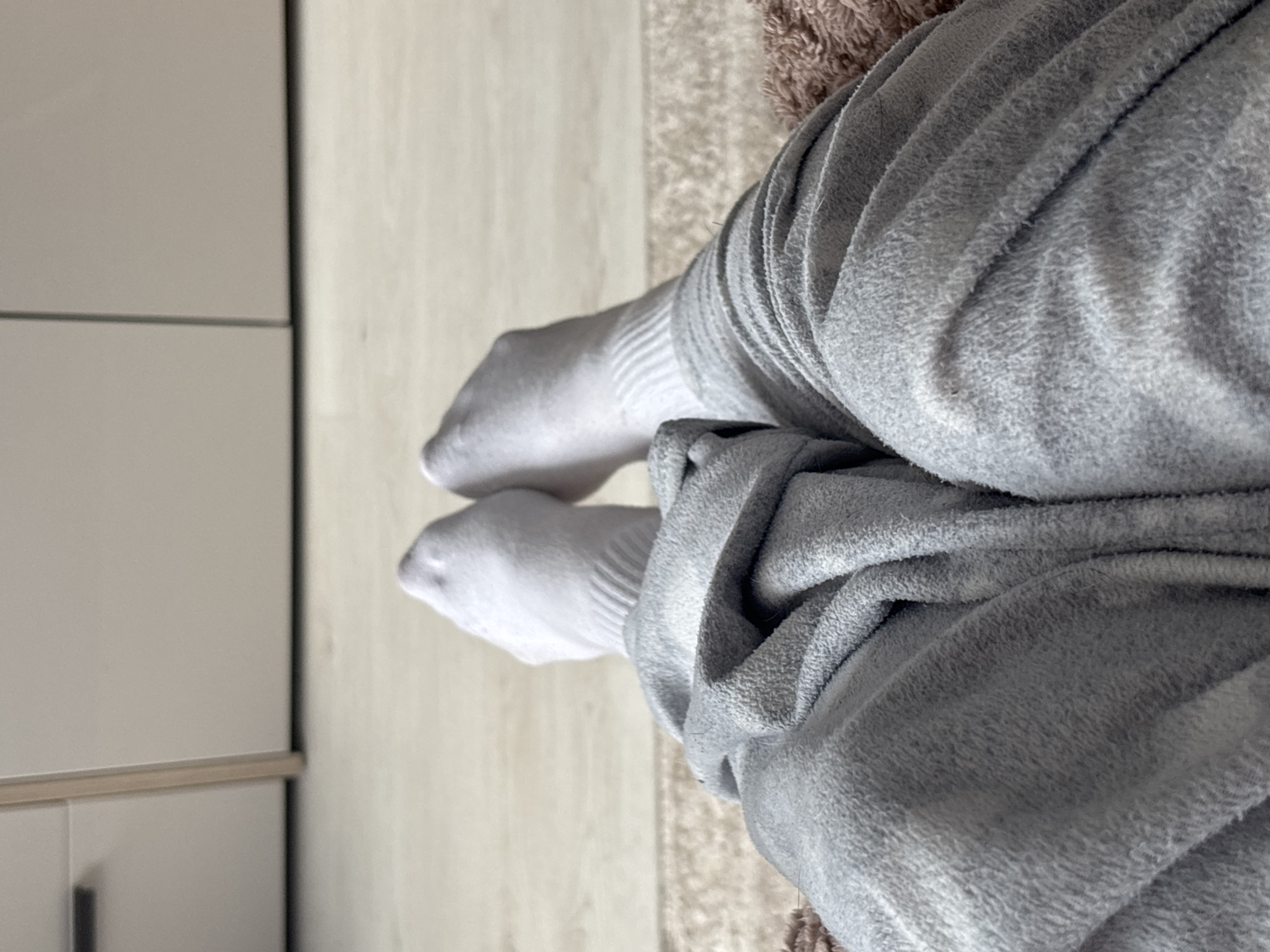 A first-person perspective shot shows a pair of feet wearing white socks, with legs draped in soft gray sweatpants. The subject is lying down on a carpeted surface, with a wooden floor and a closet visible in the background. The lighting is natural and soft, casting gentle shadows.