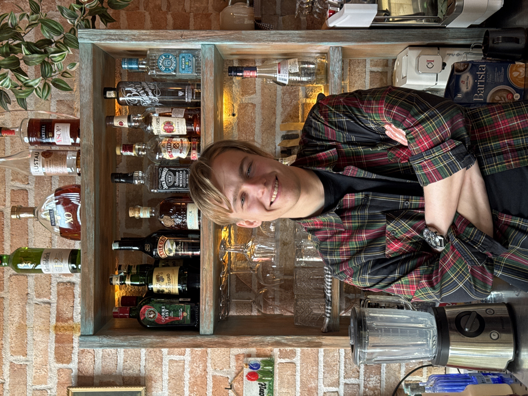 A young man with blonde hair and a wide smile is prominently featured in this image, posing with his arms crossed in what appears to be a bar setting in Rafailovići, Montenegro. He wears a plaid shirt in shades of red, green, and black over a black t-shirt, and a watch is visible on his left wrist.
The background reveals a rustic interior with exposed brick walls. Behind the man, a wooden shelving unit displays a variety of alcoholic beverages. Notable bottles include Olmecca Tequila Silver, Bols, Four Roses Bourbon, Chivas Regal XII, Jack Daniel's Tennessee Whiskey, Martell VSOP, Lustau, Glenfiddich, Martell XO, Baileys Original, Montenegro, and Gorki List.
To the man's right, a coffee machine branded "caffe d'italia" is visible, along with a "Barista" carton that reads "W LEKARA SARAC". A Philips blender sits on the counter in the foreground. A lush green plant cascades from the upper left corner, adding a touch of nature to the scene. The lighting suggests an indoor daytime setting or a brightly lit evening. The man's smiling expression conveys a friendly and welcoming atmosphere.