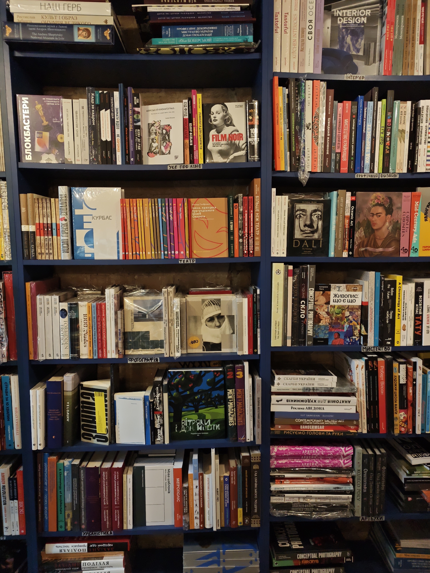 The image shows multiple shelves of books in what appears to be a bookstore or library. The books are of various sizes, colors, and languages, with many featuring Ukrainian titles and authors. The shelving unit is dark blue.
On the top shelf, there are books with titles such as "НАШ ГЕРБ", "ЛЮСТРАЦІЯ ВЕЛИКОГО", "The Andrey Sheptytsky National Museum in Lviv", and "INTERIOR DESIGN".
The second shelf from the top displays books like "БЛОКБАСТЕРИ", "Кіно", "FILM NOIR", and "100 MOVIES".
The third shelf contains books with titles including "КУРБАС", "ТЕАТР", and various art and photography books.
The fourth shelf features more books with titles such as "ФОТОГРАФІЯ", "DALI", and a book with a portrait of Frida Kahlo.
The fifth shelf from the top shows books with titles like "ВІТРАКИ І ДОЩ", "МЕТРОПОЛІС", and "URBANISTIKA".
The bottom shelves visible contain more books, including ones with titles related to "СКАРБИ УКРАЇНИ", "CONCEPTUAL PHOTOGRAPHY", and various other literary works.
The overall impression is a well-stocked collection of books, likely a retail or public space dedicated to literature and art. The style of the books and the presence of Ukrainian text suggest a location within Ukraine, possibly Kyiv.