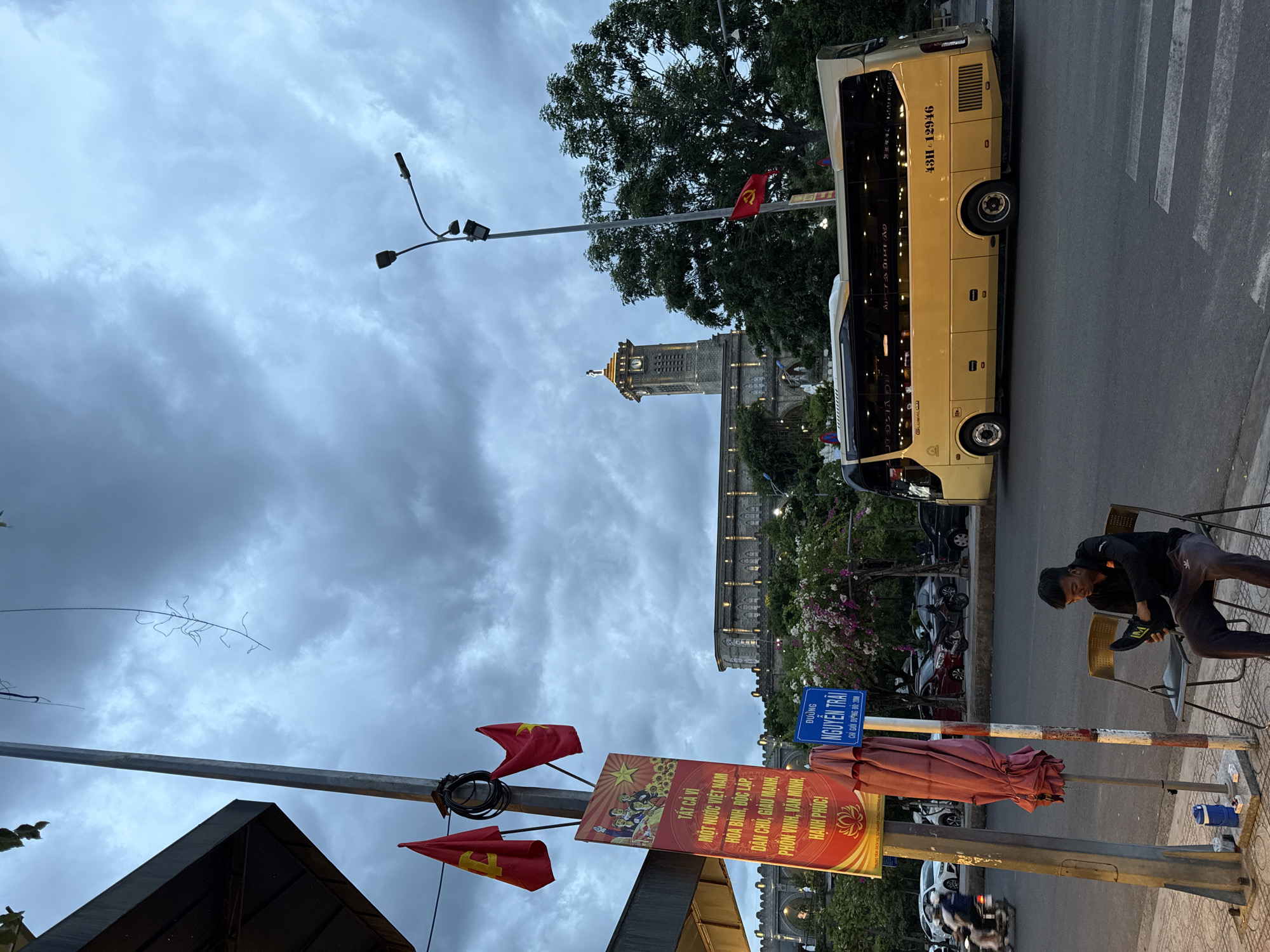 The image depicts a street scene in Nha Trang, Vietnam, at dusk. A prominent yellow tour bus with license plate 43H-129.46 is parked on the right side of the road. In the foreground, a young man in dark clothing is sitting on a folding chair, holding a black shoe, seemingly putting it on. To his left, a blue thermos and some small items are placed on the sidewalk next to a utility pole adorned with two red Vietnamese flags and a rectangular banner with Vietnamese text and images of people. A large, folded pink umbrella is leaning against the utility pole. Across the street, a grand stone building with a clock tower and arched windows stands out against the overcast sky. Trees and flowering bushes line the area in front of the building. In the background, the street is busy with a few cars and motorcycles. The overall atmosphere is calm, with the sky showing signs of an impending twilight or possibly rain. The location appears to be a public area, possibly near a tourist attraction or transportation hub.