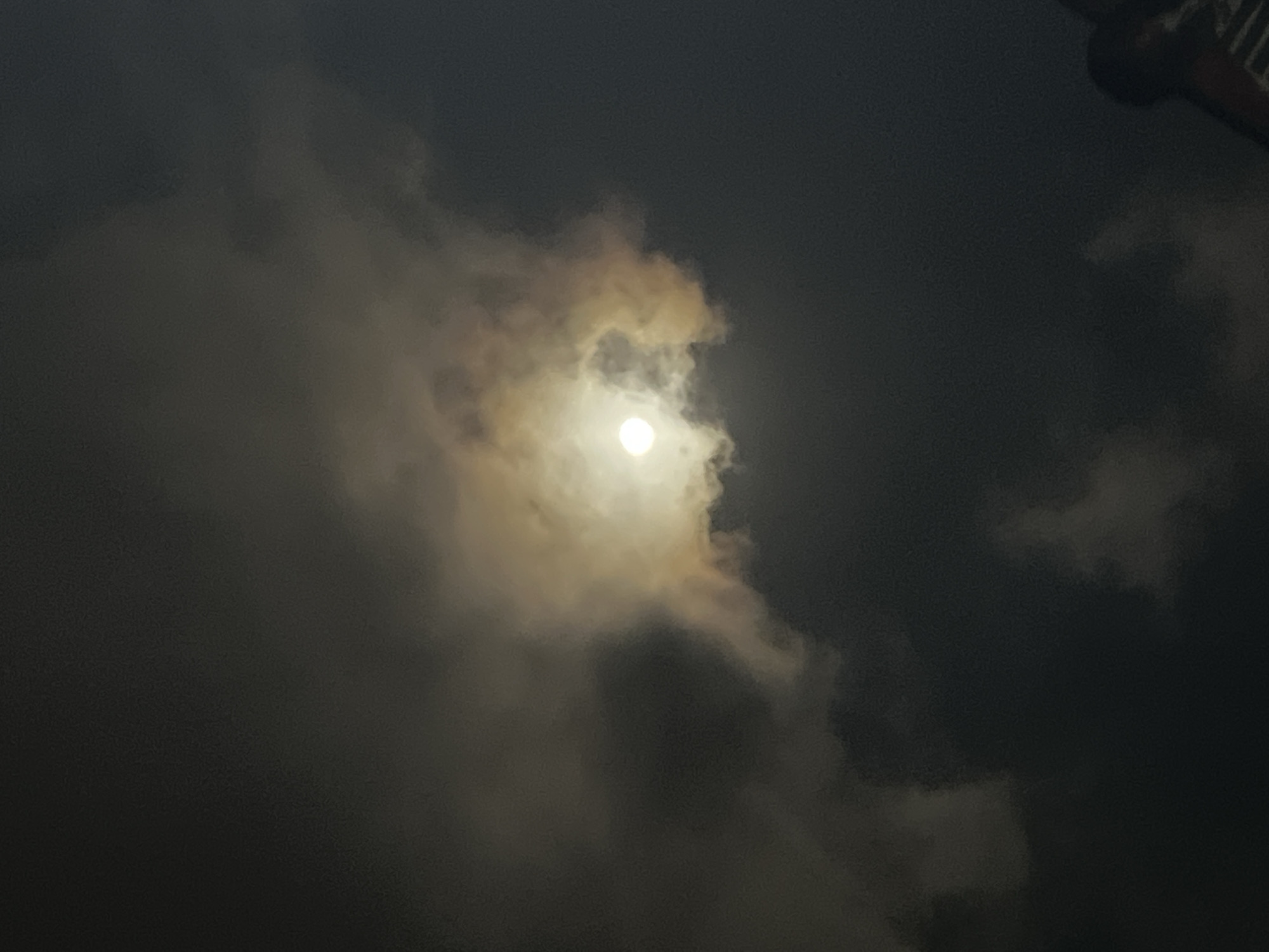 A full moon is partially obscured by clouds, creating a luminous halo effect. The sky is dark, indicating nighttime, and the clouds are a mix of dark grays and lighter, yellowish-white hues illuminated by the moon. The image appears to be taken from a low angle, looking upwards. In the bottom right corner, the silhouette of a building's railing and roofline is visible, suggesting an urban environment. The scene is devoid of any people or active subjects. The image captures a moment of a moonlit night with atmospheric cloud cover over the city of Denpasar, Indonesia.