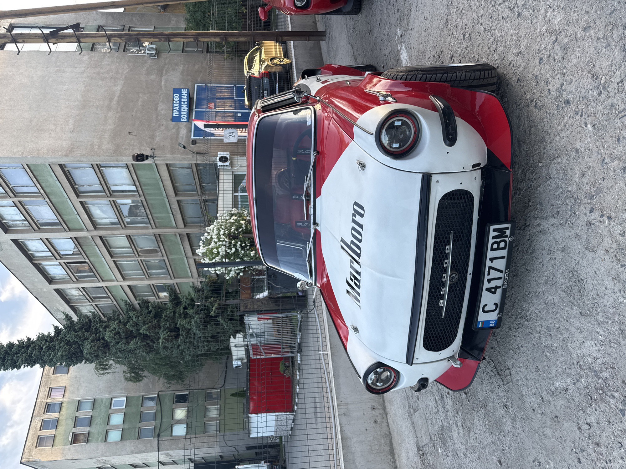 In the city of Busmantsi, Bulgaria, a modified vintage Skoda car, painted in a red and white Marlboro livery, is parked on a paved surface. The car features wide fender flares and aftermarket headlights. The vehicle's license plate reads "C 4171 BM" and displays the "SKODA" logo. Behind the car, a chain-link fence encloses an area with red industrial doors and some greenery. Further in the background, a multi-story Brutalist-style building with green accents dominates the scene, and a tall evergreen tree stands prominently in front of it. A sign with blue lettering reading "ПРАХОВО БОЯДИСВАНЕ" (Powder Coating) is visible on the building. The sky above is overcast.