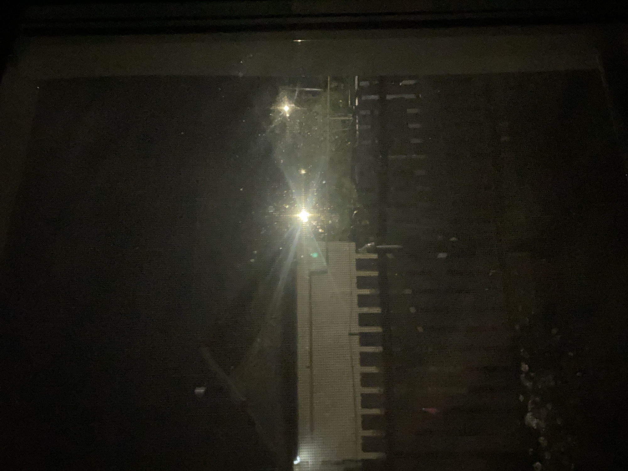 The image is a dark, grainy photograph taken at night through a window, likely from inside a residence. The view outside shows a dimly lit outdoor scene. In the middle ground, there is a light-colored building with a prominent dark, sloped roofline. The building appears to have a row of evenly spaced, dark rectangular openings along its lower facade, suggesting a structure with an arcade or enclosed walkway. In front of this building, there is a dark wooden fence, also with regularly spaced vertical elements. Beyond the fence, trees and vegetation are visible, illuminated by two bright, diffused light sources that appear to be streetlights or security lights. The lights create a lens flare effect and scatter light, contributing to the overall haziness of the image. There are small, bright specks scattered throughout the scene, possibly dust or water droplets on the windowpane or in the air. The foreground is very dark, with some indistinct white or light-colored debris on the ground, and a faint pink object that may be a flag or marker. The overall impression is of a suburban or semi-rural environment at night, with artificial lighting casting a glow on the surroundings. The mention of Shiryaevo, Russia, provides a geographical context for the scene, suggesting a location in that specific region.