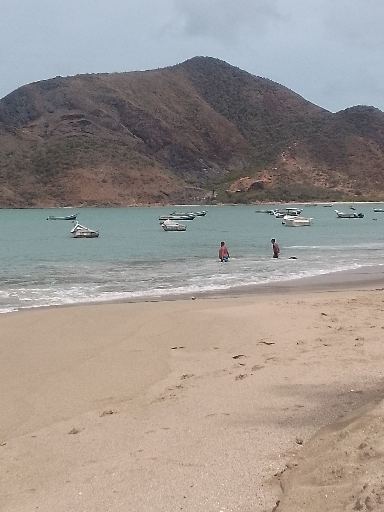 The image shows a beach in Juan Griego, Venezuela, with a large mountain in the background. Two people are wading in the water. Several boats are anchored in the bay. The beach has light-colored sand with visible footprints. The sky is overcast.