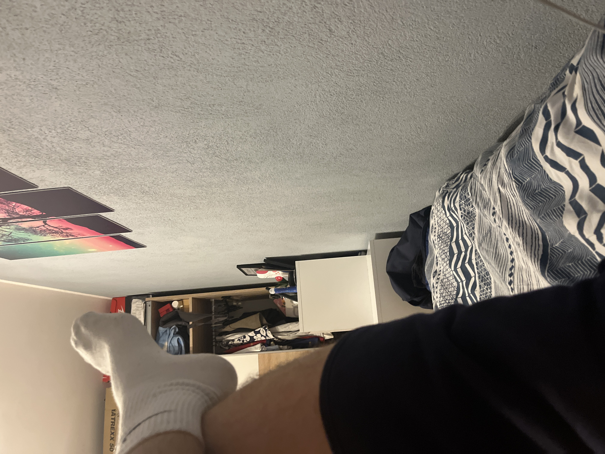The image shows a domestic interior with a partial view of a person. The person's leg, covered in a white sock, is prominent in the foreground, alongside the dark fabric of their clothing. Behind the leg, a white dresser with open drawers is visible, revealing stored clothes and accessories. A bed with a blue and white patterned duvet occupies the upper right. On the left, there are framed artworks hanging on the wall, near a wall corner. The ceiling has a textured surface. It is not possible to determine the location in Pokrovnik, Bulgaria from the image alone.