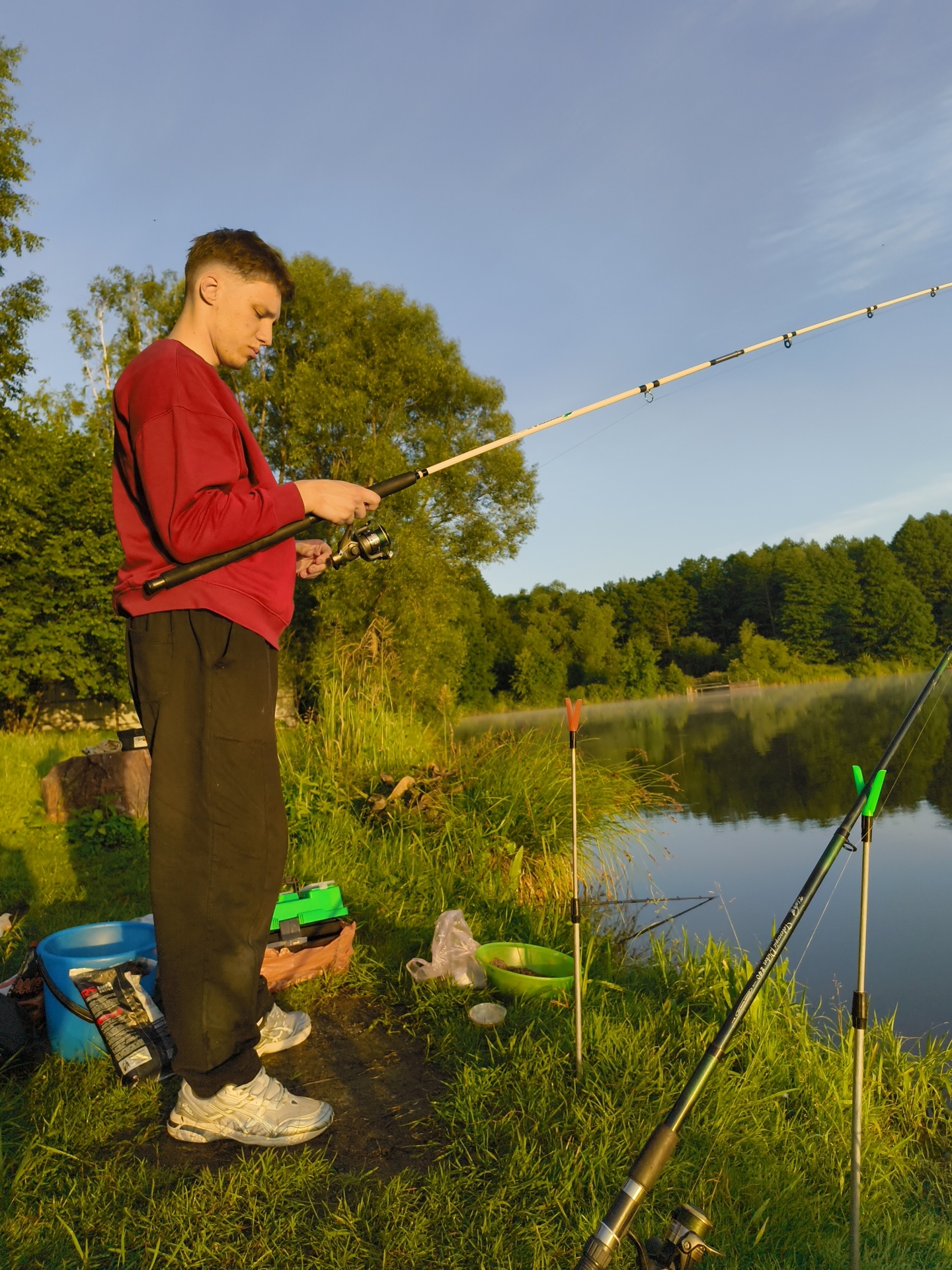 A young man stands on the grassy bank of a lake, holding a fishing rod. He is wearing a maroon sweatshirt and black pants. The lake is calm, with trees lining the opposite shore. Several other fishing rods are set up nearby. A blue bucket and a green tackle box are on the ground next to him. The sky is clear and blue, with the sun casting long shadows. The scene appears to be a peaceful, outdoor setting.