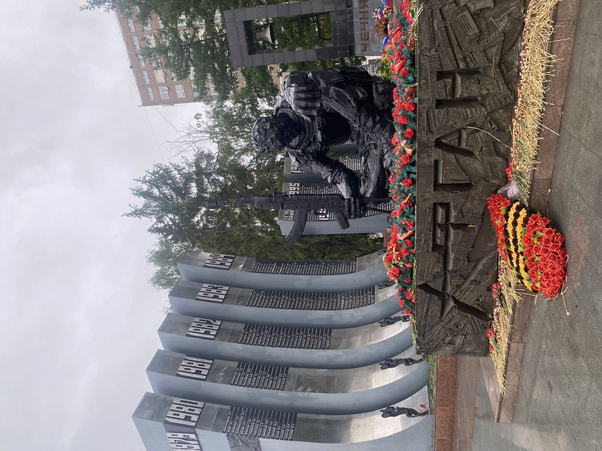The image shows a war memorial in Yekaterinburg, Russia, dedicated to soldiers who died in Afghanistan and Chechnya. A bronze statue of a soldier kneeling with a rifle is in the center of the frame. The memorial has curved walls with names listed on them. The wall segments are marked with years, including 1980 and 1985. Red and yellow floral wreaths are placed at the base of the statue. A building can be seen in the background through the trees. The sky is overcast.