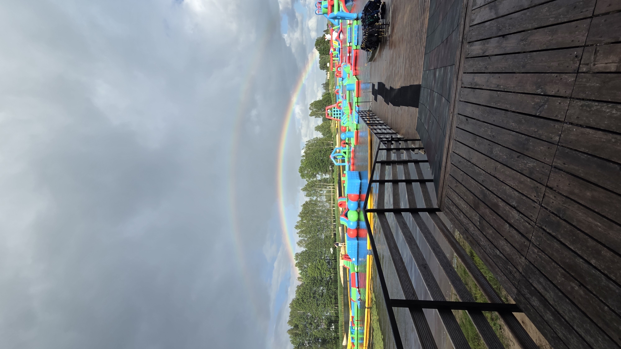 The image captures an outdoor water park located in Jaunmārupe, Latvia. A prominent double rainbow arches across a cloudy sky, with the main rainbow framing a vibrant collection of inflatable water attractions floating on a body of water. These colorful structures, including slides, climbing walls, and various shapes, are scattered across the water's surface. The park appears to be set against a backdrop of trees and a distant shoreline. In the foreground, a wooden deck with a dark railing leads towards the water, providing a vantage point of the park. There are a few individuals visible, some on the inflatable attractions and others near the edge of the water, suggesting the park is operational. The overall scene depicts a recreational area on a potentially overcast day with a striking natural phenomenon.