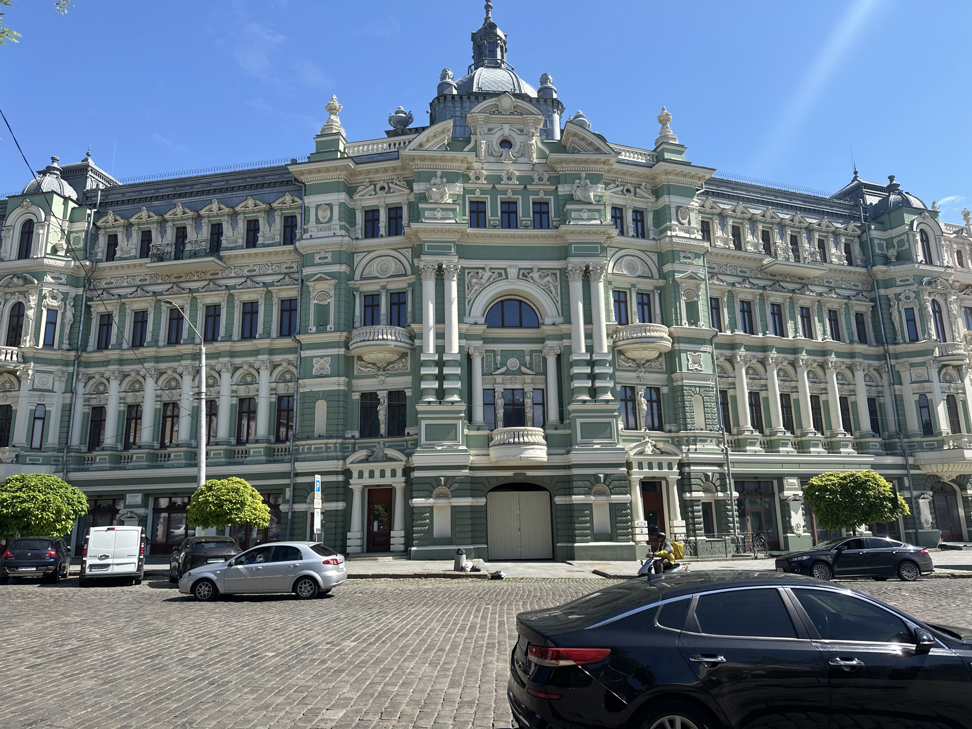 The image depicts a large, ornate building with a light green and white facade in an urban environment. The building has multiple stories, numerous windows, columns, balconies, and decorative elements. At the top, there is a dome-like structure.
In the foreground, a cobblestone street is visible with several cars parked along the side. A black car is positioned closest to the viewer, partially obscuring the view. A silver car and a white van are parked further down the street, along with a black sedan. A person on a moped is on the sidewalk. Trees with rounded foliage are situated along the sidewalk. The sky is clear and blue.