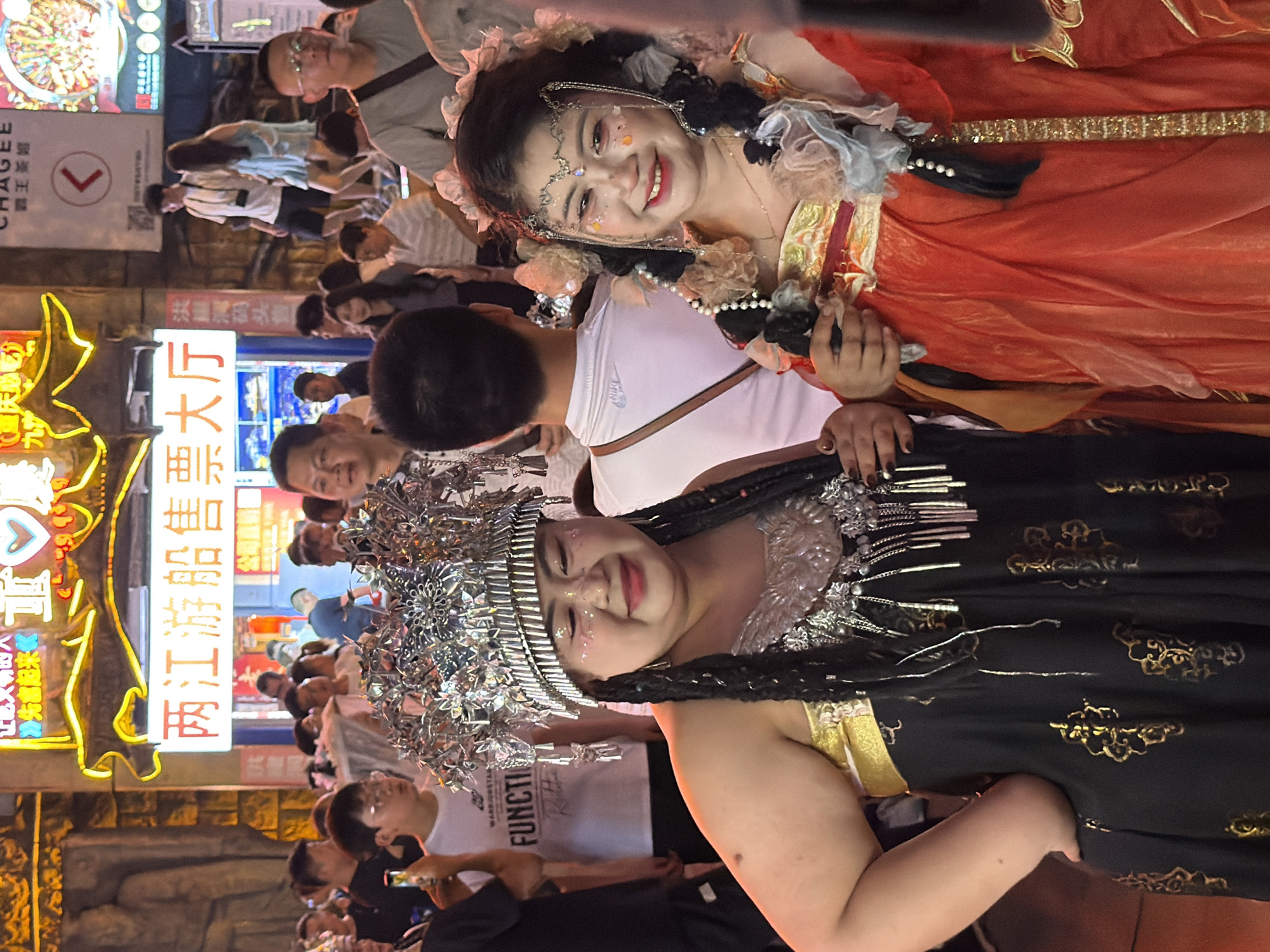 This is a medium shot photograph taken at night in Yuzhong District, China, likely during a festival or event. The foreground is dominated by two women in elaborate costumes. The woman on the left wears a black and gold dress with a highly ornate, silver headdress adorned with tassels and intricate patterns. Her makeup includes glitter on her cheeks and eyes, and she has dark braided hair. To her right, another woman is dressed in a vibrant orange and gold outfit, also with a decorative headdress featuring flowers and pearls, and similar glittery makeup. Both women are smiling and appear to be posing for the camera.
Behind them, a crowd of people are visible, many of whom are looking towards the foreground or the street. The background features illuminated signage and buildings, indicative of a commercial or entertainment district. One sign in Chinese characters appears to advertise a ticket office. The overall atmosphere is lively and festive, suggested by the neon lights and the presence of costumed performers. The lighting is artificial and predominantly warm, casting a glow on the subjects and the surroundings.