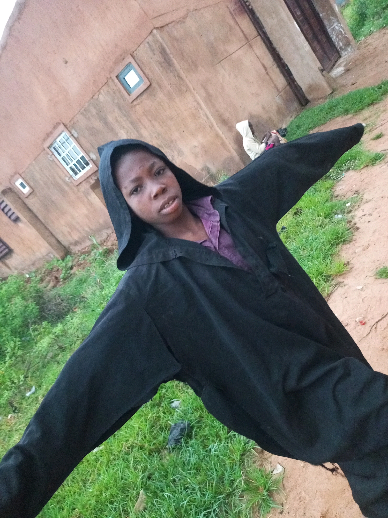 A young person wearing a black hooded robe over a purple shirt stands in front of a two-story building with a sandy ground and sparse green grass. The building's facade is a reddish-brown color, with visible windows. The person's arms are outstretched to the sides. In the background, another figure in a light-colored hooded garment is partially visible, sitting on the ground. The scene appears to be outdoors in a casual, possibly rural or suburban environment within Tempure, Nigeria.