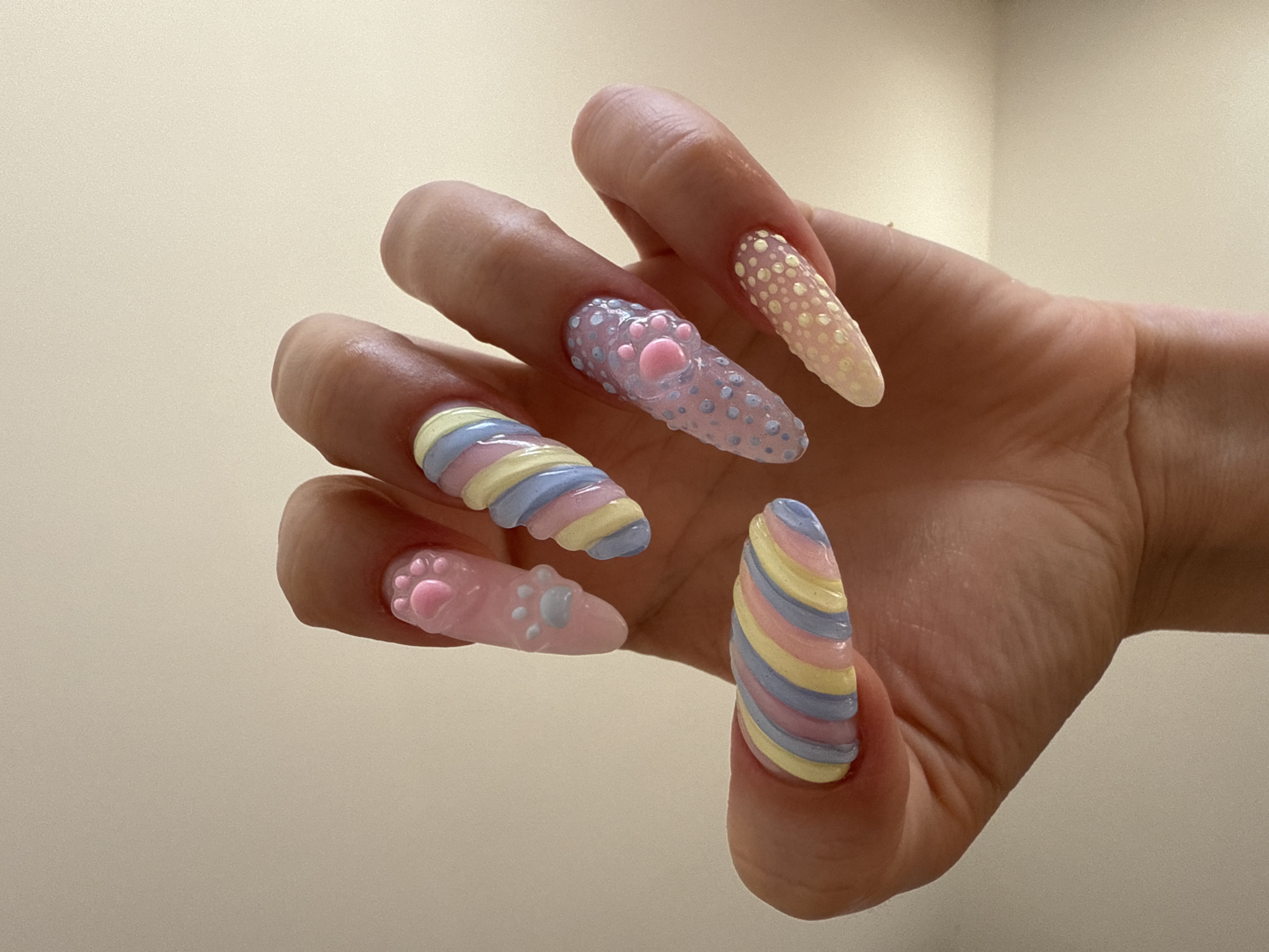 A person's hand with long, decorated fingernails is displayed against a light-beige background. The nails are adorned with pastel colors and various patterns, including a swirled candy-stripe design, raised dots, and paw prints. The composition focuses on the hand and its intricate nail art.