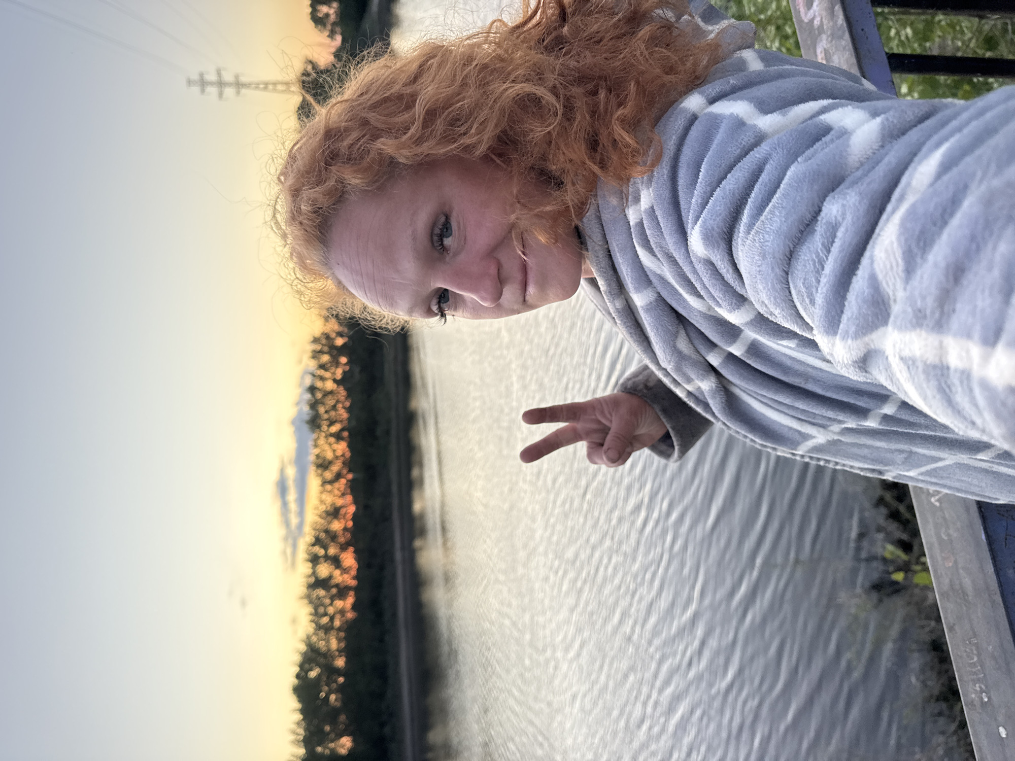 An outdoor, medium-shot selfie shows a woman with curly red hair looking at the camera. She is wearing a grey and white checkered robe. Her left hand is raised, forming a peace sign, in front of a body of water that reflects the orange and yellow sky of a sunset. A line of trees is visible on the far bank of the water, with a transmission tower in the background. The woman appears to be on a wooden platform or deck overlooking the water. This scene is located on the Kama River in Chaikovsky, Russia.