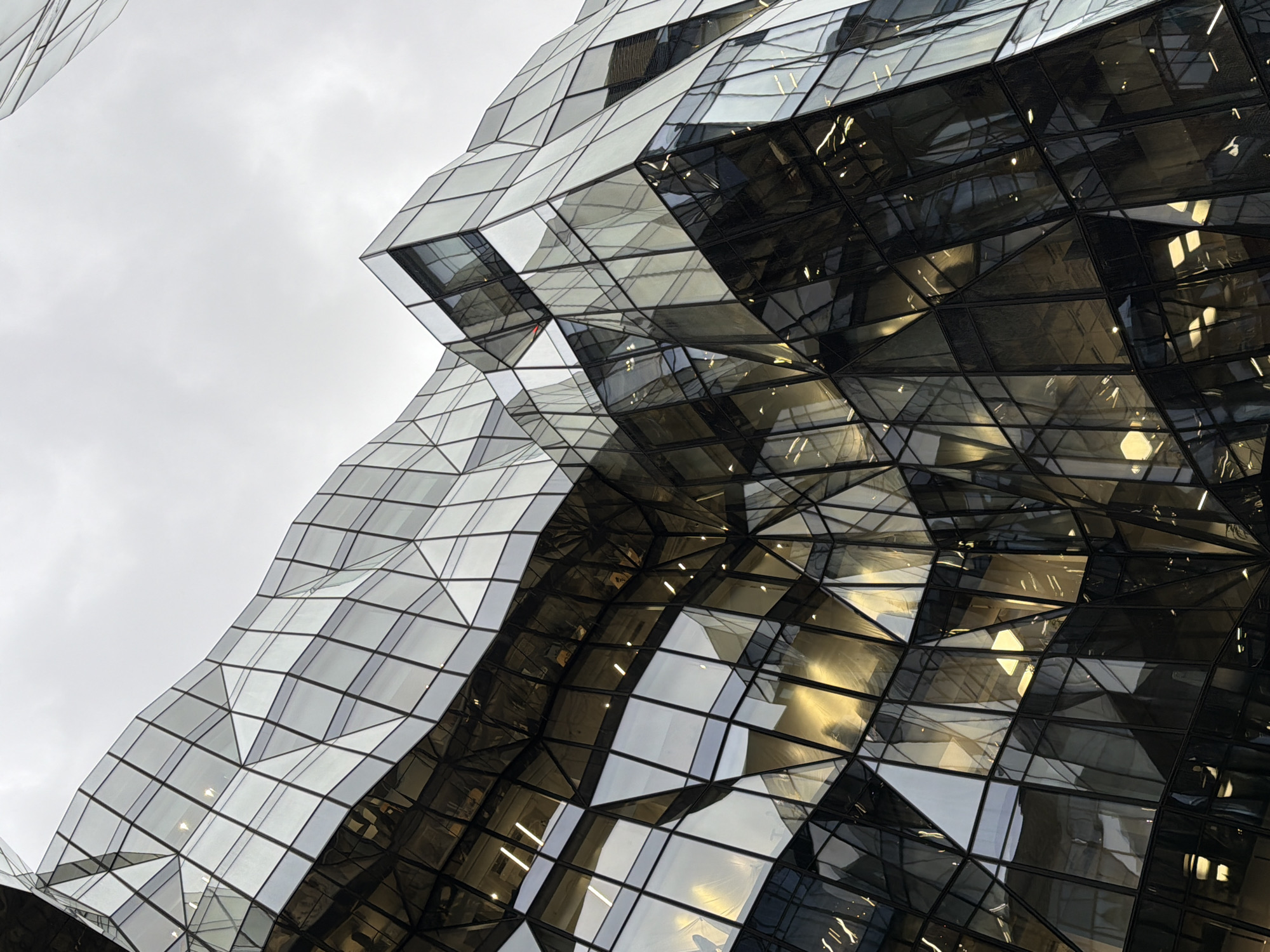 The image shows a close-up of a building facade with a geometric, tessellated design. The facade is predominantly made of glass panels, reflecting both external light from an overcast sky and internal illumination. The reflective properties of the glass create contrasting tones, with some panels appearing bright and others dark. The building is located in the city of Unknown, Unknown. The architecture is modern and angular. There are no people visible in the image.