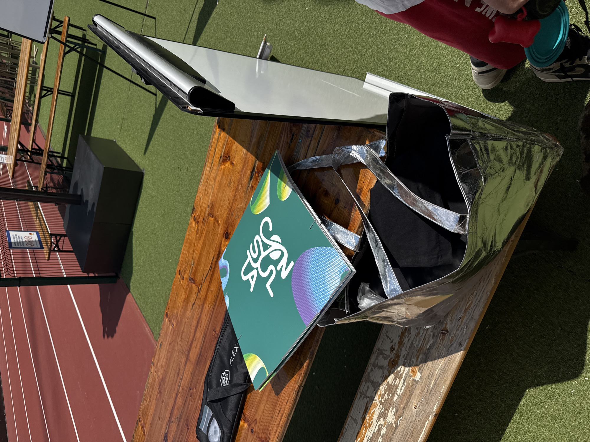 This image captures an outdoor scene at a sports facility in Milan, Italy, on a sunny day. A worn wooden picnic table, with peeling paint on its bench, occupies the foreground. On the table, from left to right, are a black soft case with "FLEX" and a geometric logo printed in white, a closed green booklet with a stylized white script design (partially obscured) and abstract purple, yellow, and white circular patterns, and a metallic silver tote bag with dark interior lining.
To the right of the table, the lower half of a person is visible, wearing red athletic pants, white socks, and black and white Nike high-top sneakers. The person is holding a red pet water or treat dispenser with a teal cup. Directly behind the table, a white pull-up banner stand is visible.
The background features a red running track with white lane markers, bordered by artificial green turf. A dark green wire fence runs along the edge of the turf, behind which more wooden picnic tables and benches are arranged. A large black square base, likely for a parasol or display, is situated on the turf. Strong shadows are cast across the ground and some objects, indicating bright sunlight. A small, unreadable white sign with blue text and graphics is affixed to the fence.