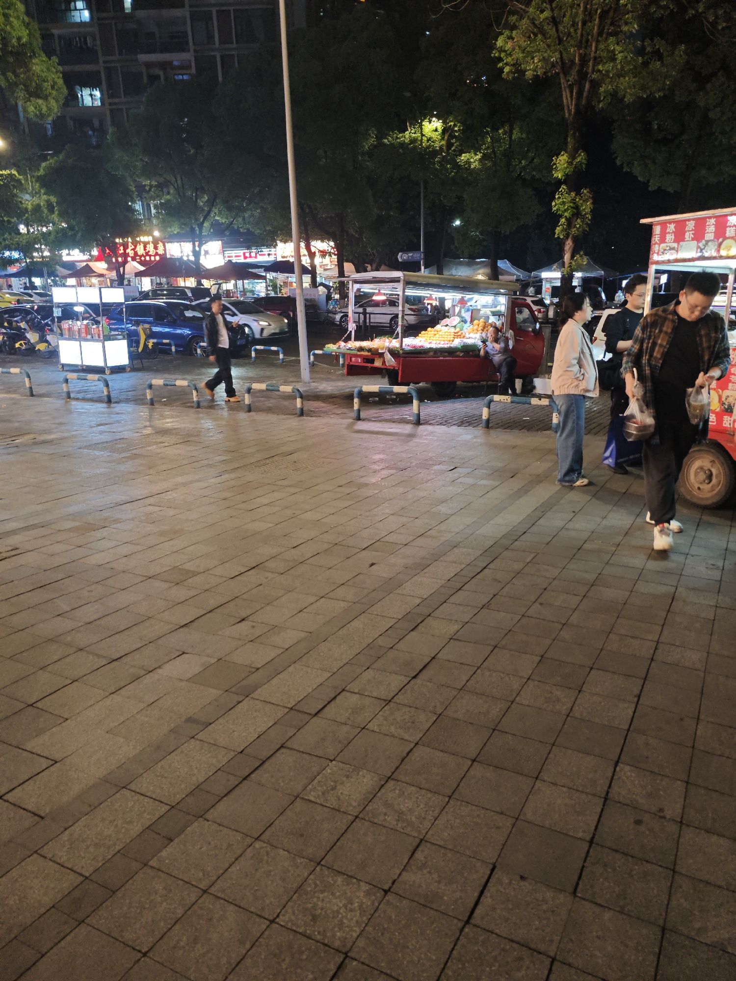 The image shows an evening street scene in Liangjiang, China, likely near a food market. The ground is paved with gray stone blocks. Several food carts are visible; one is selling fruit, and another offers sweet treats. People are milling about, including individuals buying food from the carts. Streetlights and illuminated storefronts contribute to the ambiance. Cars and motorcycles are parked on the periphery of the scene.