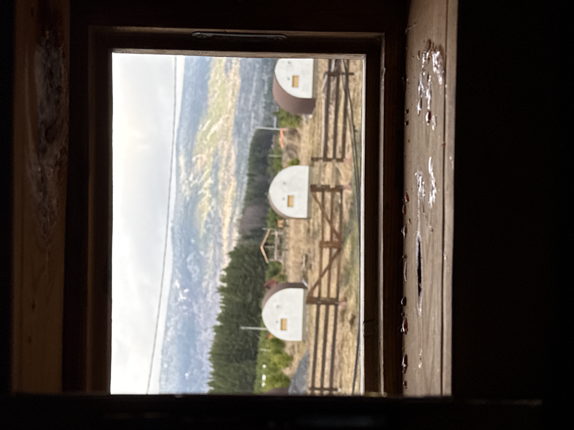 The image shows a view from inside a dark room, looking out through a wooden-framed window. The outside scene is a rural landscape featuring a hillside with a mix of trees and open grassy areas. Three small, white, semi-circular structures with brown tops and rectangular openings on their front are visible in the foreground, along with what appears to be fencing or some type of support structure. Mountains or hills rise in the background under a cloudy sky. The right side of the window frame shows signs of aging or damage. The location is Tyulyuk, Russia.