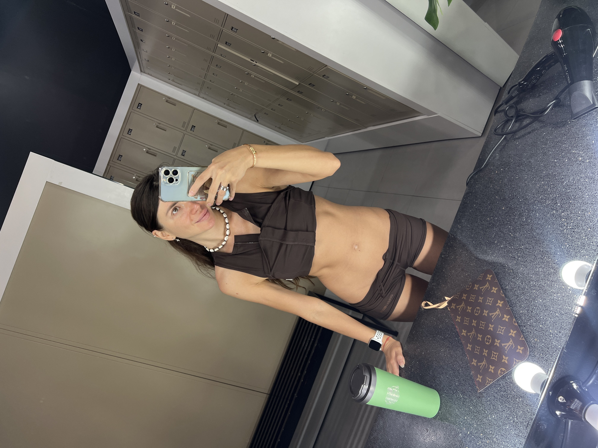 In an indoor locker room setting, a woman with long, dark hair takes a mirror selfie. She is wearing a brown athletic crop top and matching shorts, with a pearl necklace and a gold bracelet. She holds a light blue smartphone in her right hand, which has a pop socket attached. Her left hand rests on a green water bottle. In front of her is a brown Louis Vuitton pouch. The background features rows of beige lockers and a dark ceiling. The reflective surface of the locker room bench is visible in the foreground, along with a black hairdryer.