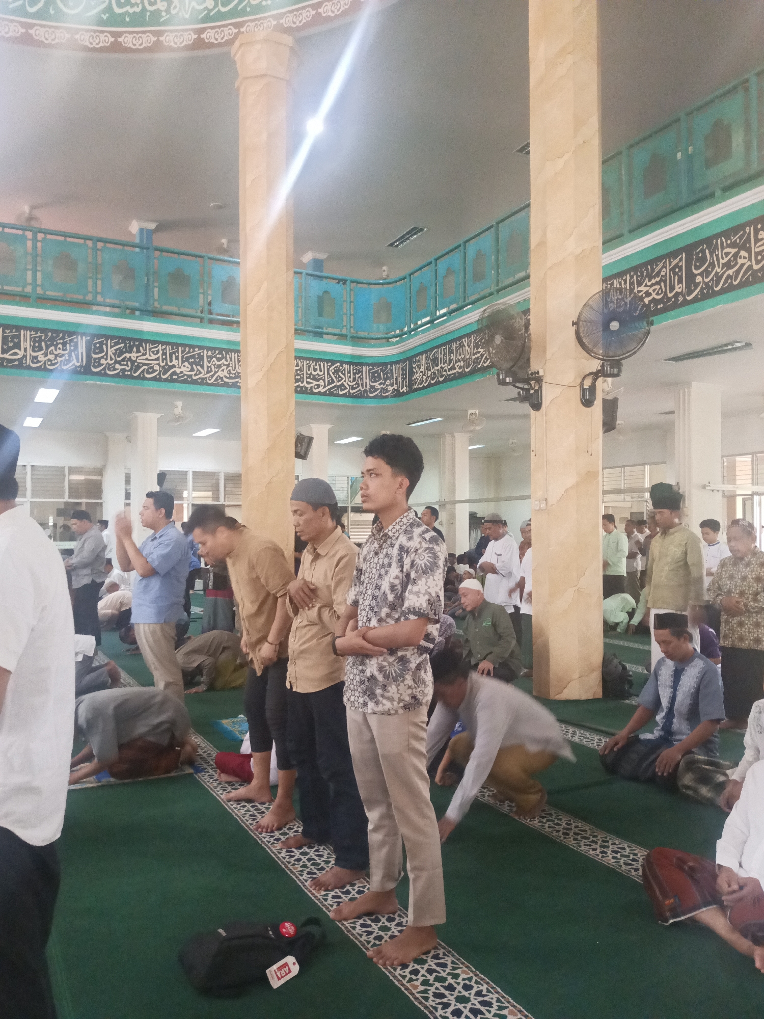The image depicts an indoor scene inside a mosque, likely in South Tangerang, Indonesia, during prayer. Several men are engaged in prayer on a green carpeted floor, with some standing and others in prostrated or seated positions. The environment features architectural elements typical of a mosque, including large, fluted columns, decorative Islamic calligraphy on the walls, and a high ceiling with lighting. Fans are mounted on the walls and columns. The individuals are dressed in a mix of traditional and casual attire, common for prayer gatherings. The overall atmosphere suggests a solemn and communal religious event.