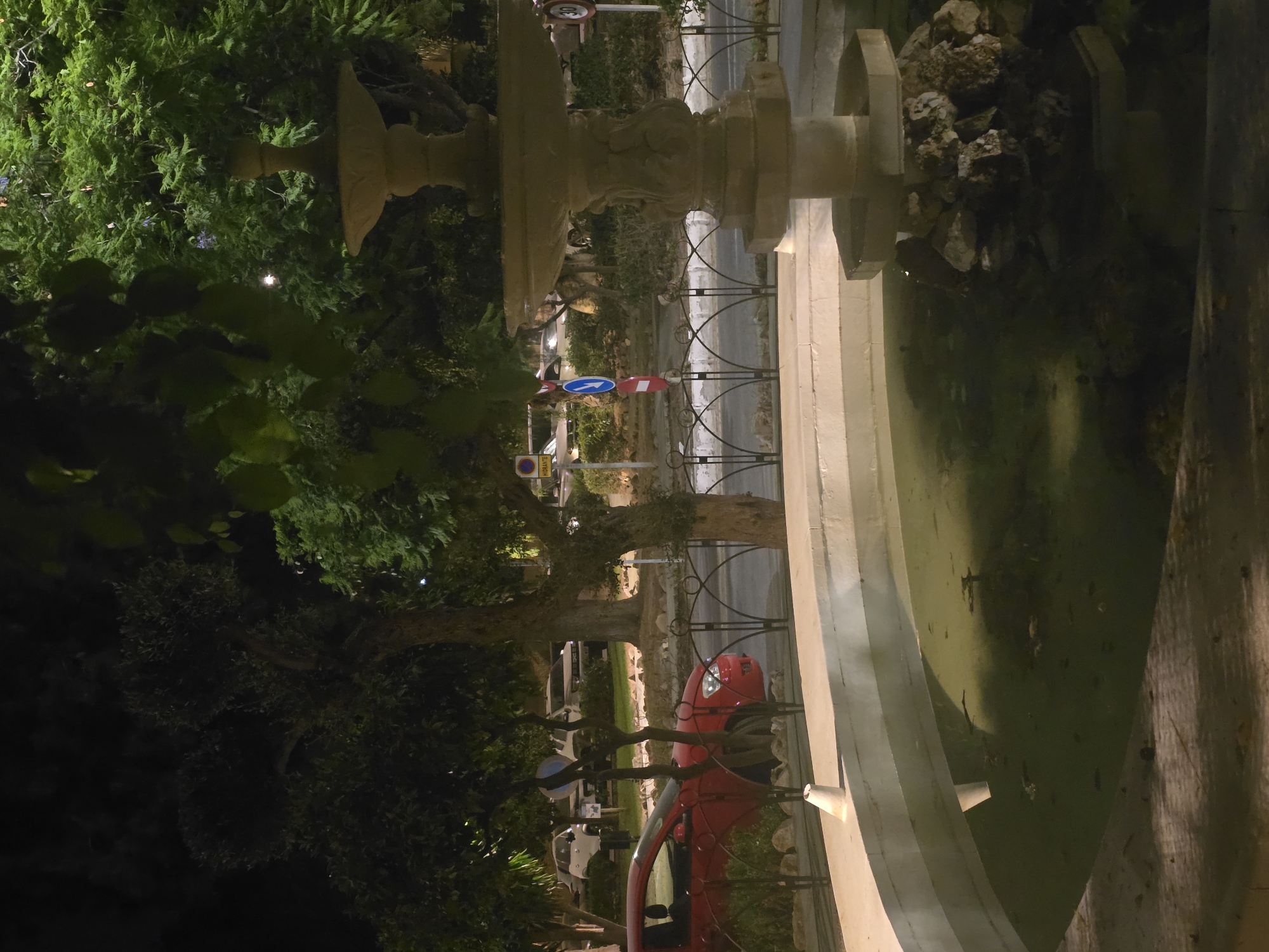 The image shows a fountain in the foreground, with a red car parked in front of it on the other side of a decorative metal fence. Behind the fence, a road is visible, with more cars and trees lining the street. The fountain appears to be made of stone and has water in its basin, which is murky and contains some debris. The scene is dimly lit, suggesting it is nighttime or early evening. The location of this fountain is Plaza de la Merced in Malaga, Spain.