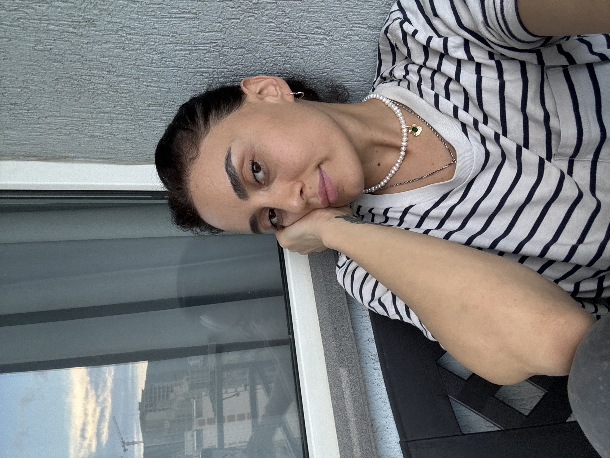 A woman is resting her head on her hand, looking towards the camera. She is wearing a black and white striped t-shirt and a pearl necklace with a green pendant. The background shows a window with a view of a city, possibly Tbilisi, Georgia, with buildings and a crane visible. The woman appears to be on a balcony or patio, with a textured wall to her right. The lighting suggests it is daytime, likely late afternoon or early evening, given the sky.